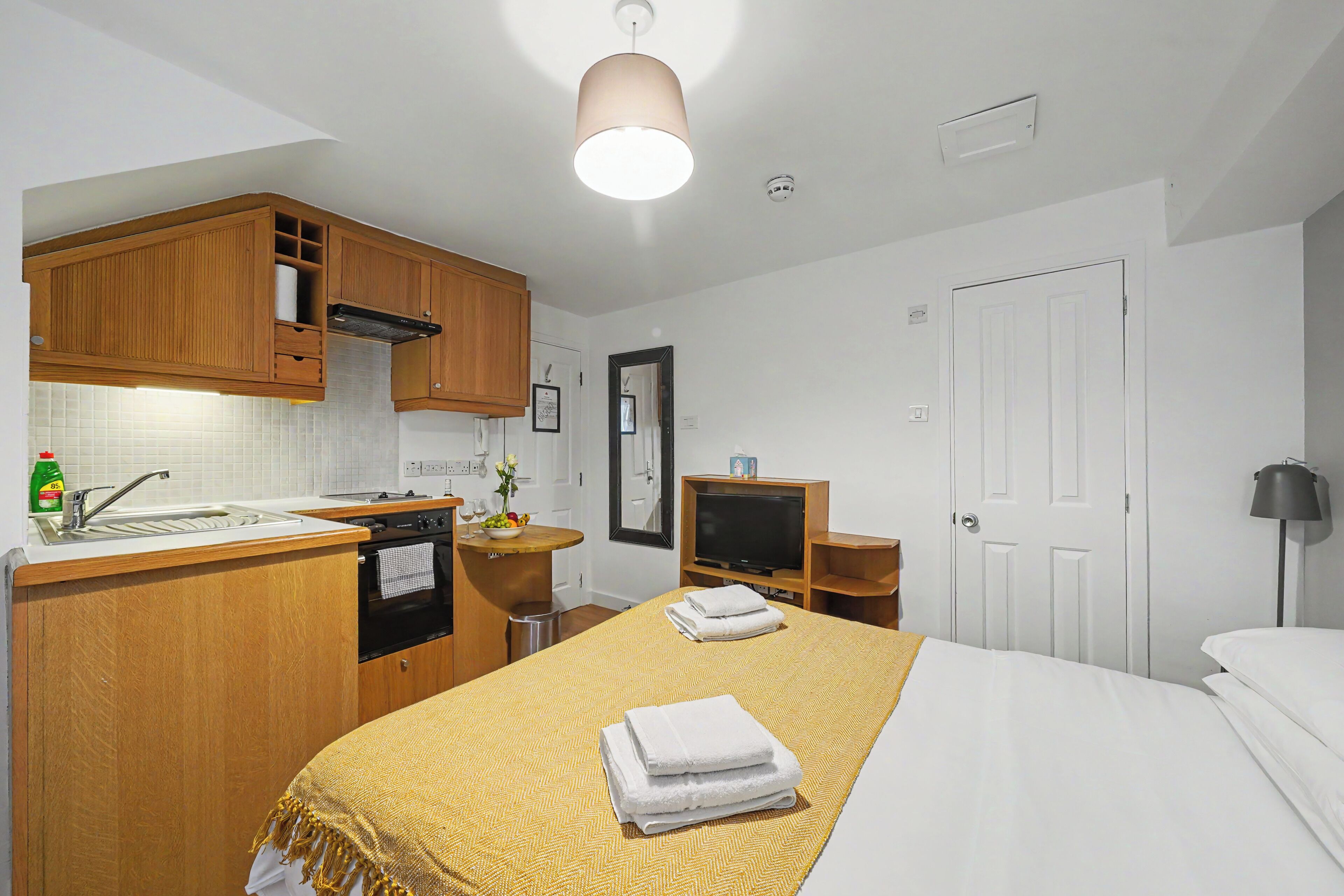 A compact hotel room with a kitchenette, a bed with a yellow patterned bedspread, and a television.