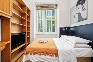 A hotel room with a bed, a television on a wooden stand, a window with a view of greenery, and a painting on the wall.