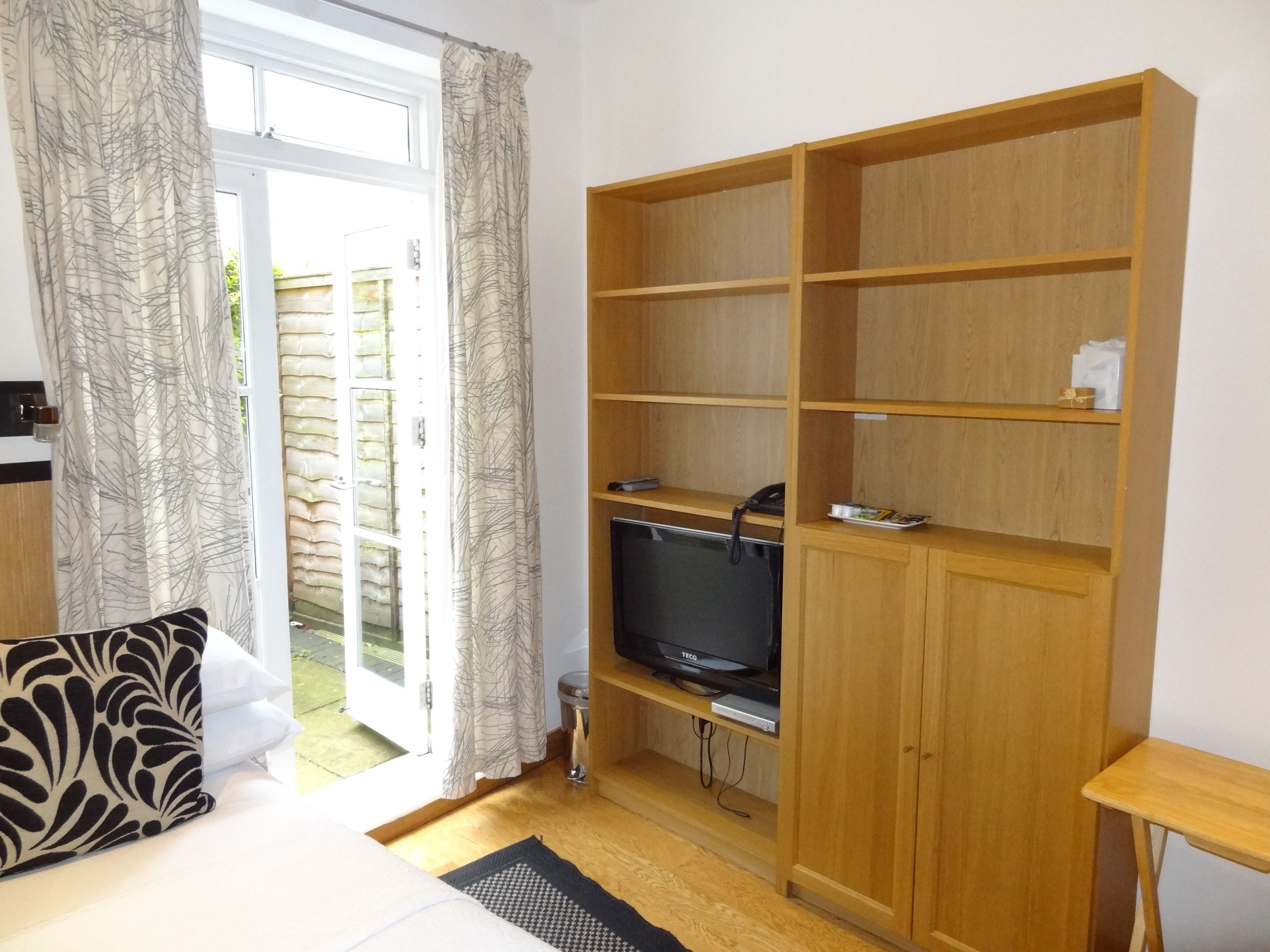 Een kamer met een bed, een televisie op een houten standaard, een houten kast en een deur naar buiten.