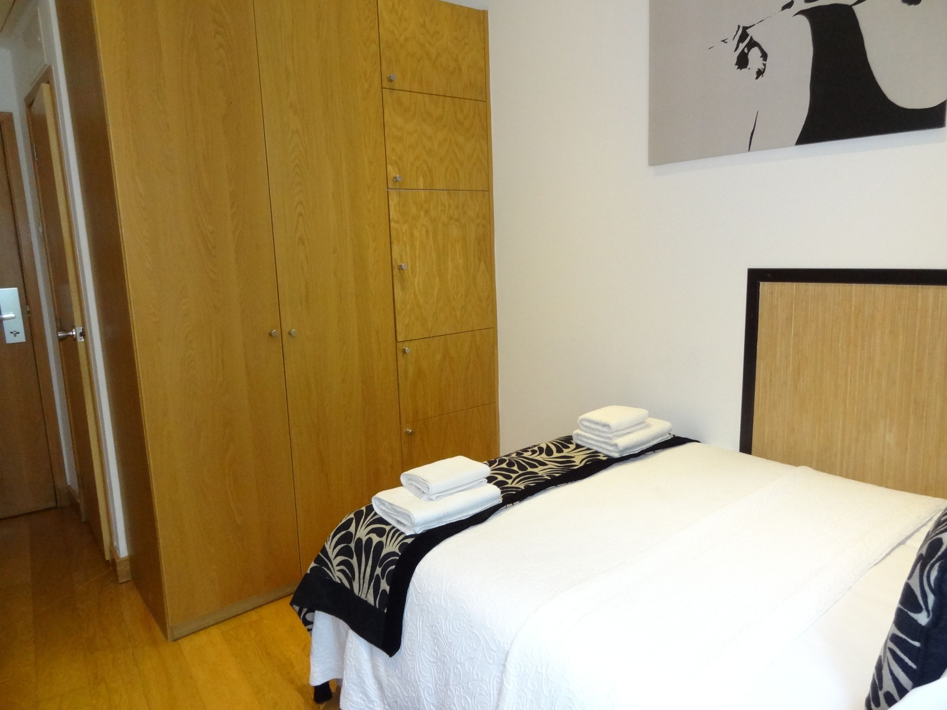 A hotel room with a wooden wardrobe, a bed with a white and black patterned bedspread, and a wall-mounted artwork of a tree.