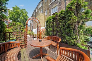 A wooden deck with a round table and benches, surrounded by greenery and brick buildings.