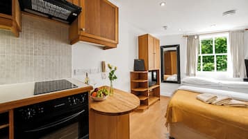 Une cuisine compacte équipée d’une cuisinière, d’une petite table à manger et d’un panier de fruits. On y trouve également un lit avec vue sur la fenêtre et un miroir.