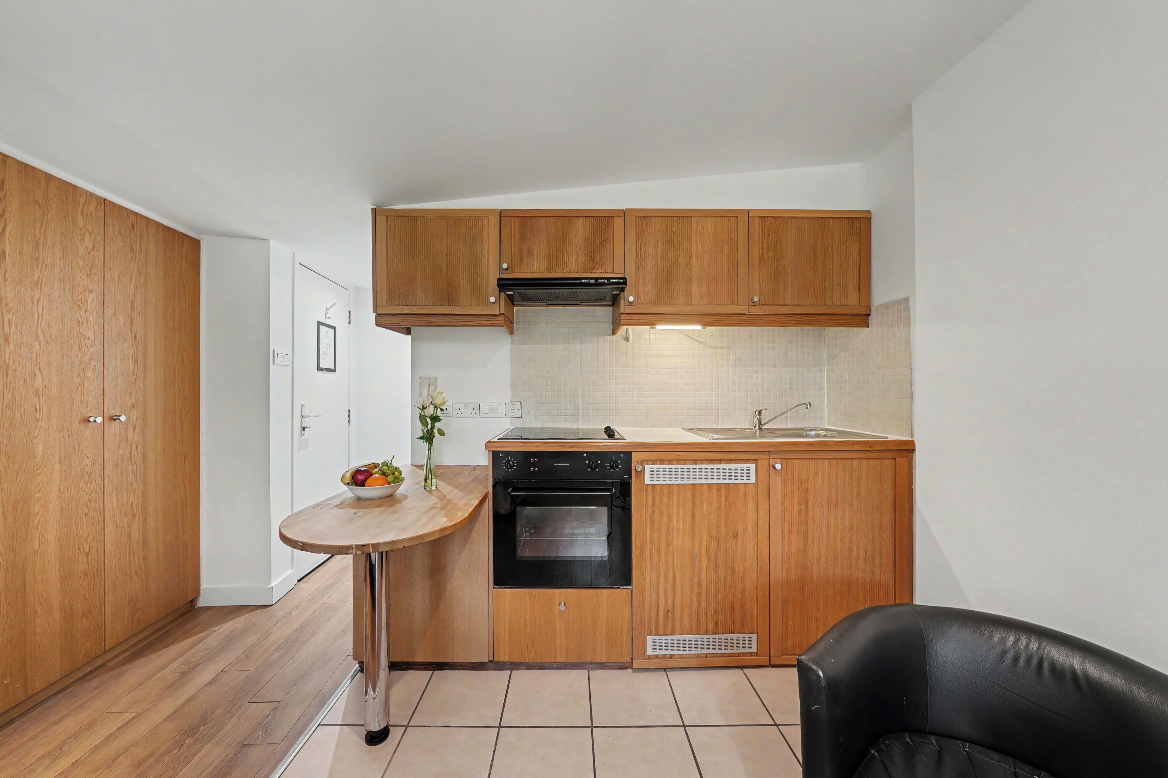 A compact kitchen with wooden cabinets, a built-in oven, a small round table with a bowl of fruit, and a black chair.