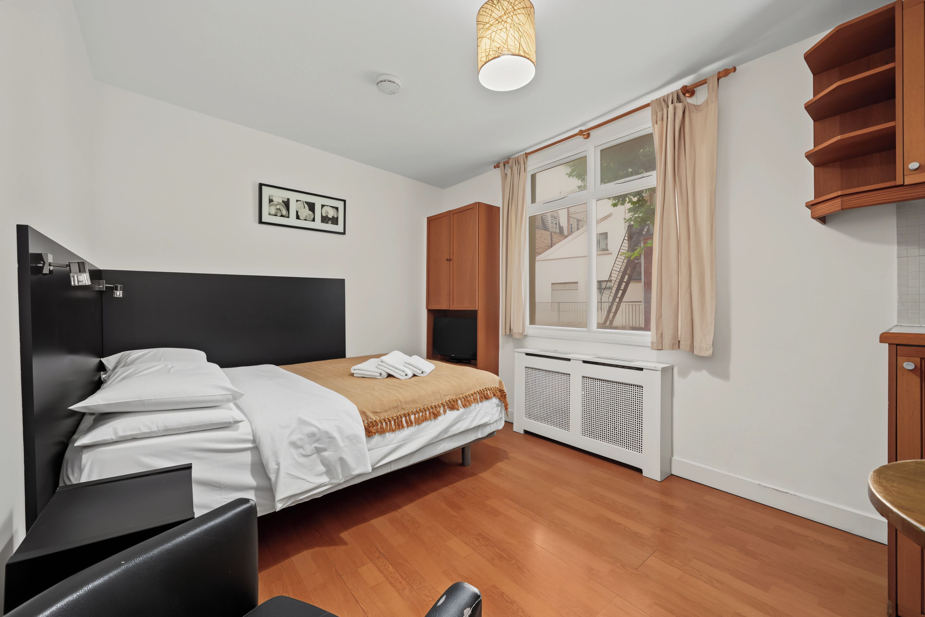 Ein Schlafzimmer mit Bett, Schreibtisch, Kleiderschrank, Heizkörper und einem Fenster mit Vorhängen.