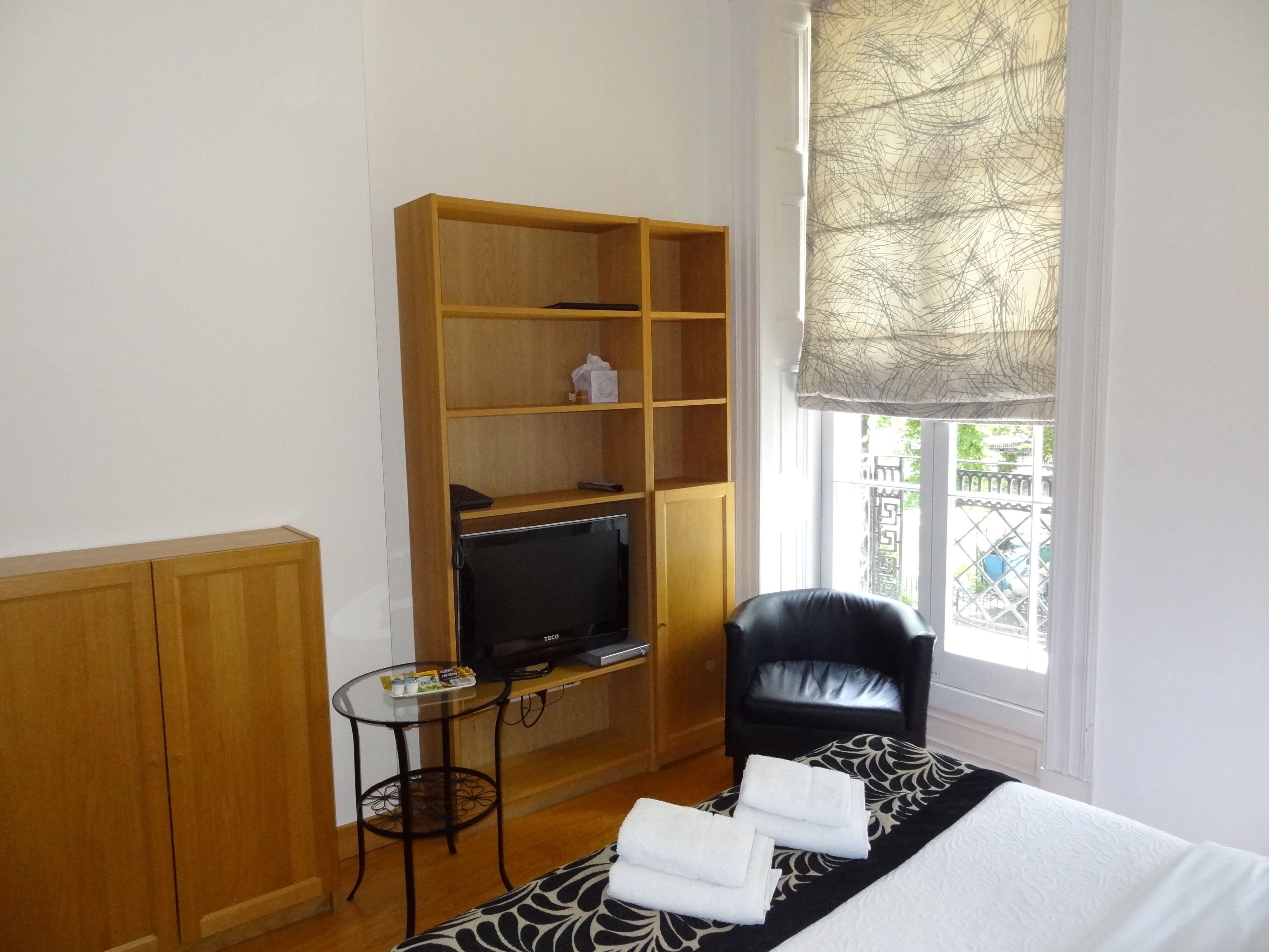 A compact room with a bed, a television, a small table, a chair, and a window with a view of a gate and greenery.