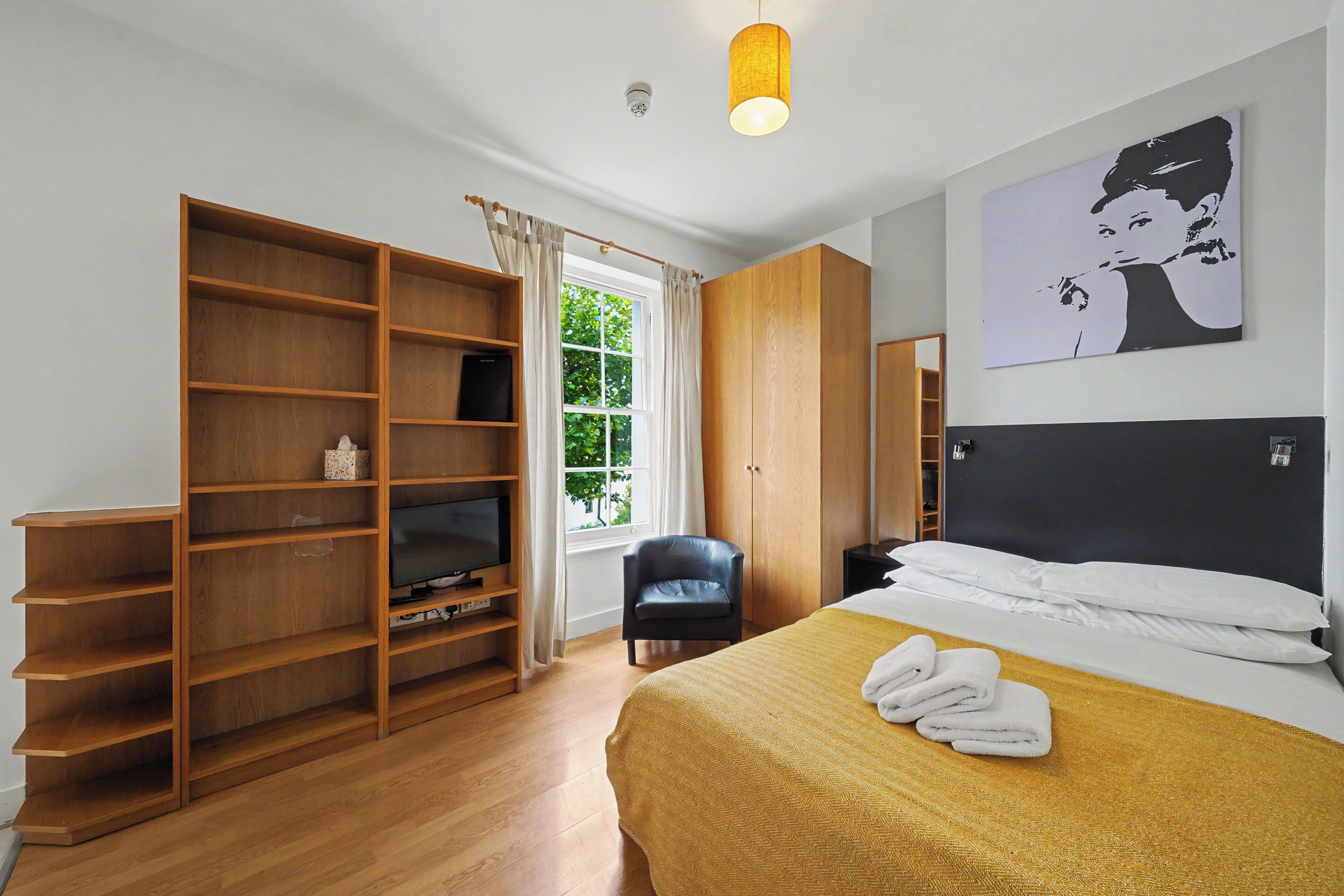 Habitación de hotel con cama, televisor sobre una estantería de madera, armario, una silla y una ventana con cortinas.