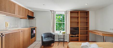 A compact kitchen with wooden cabinets, a dining area with a table and chairs, a bed with a blanket, and a window with a view of greenery.
