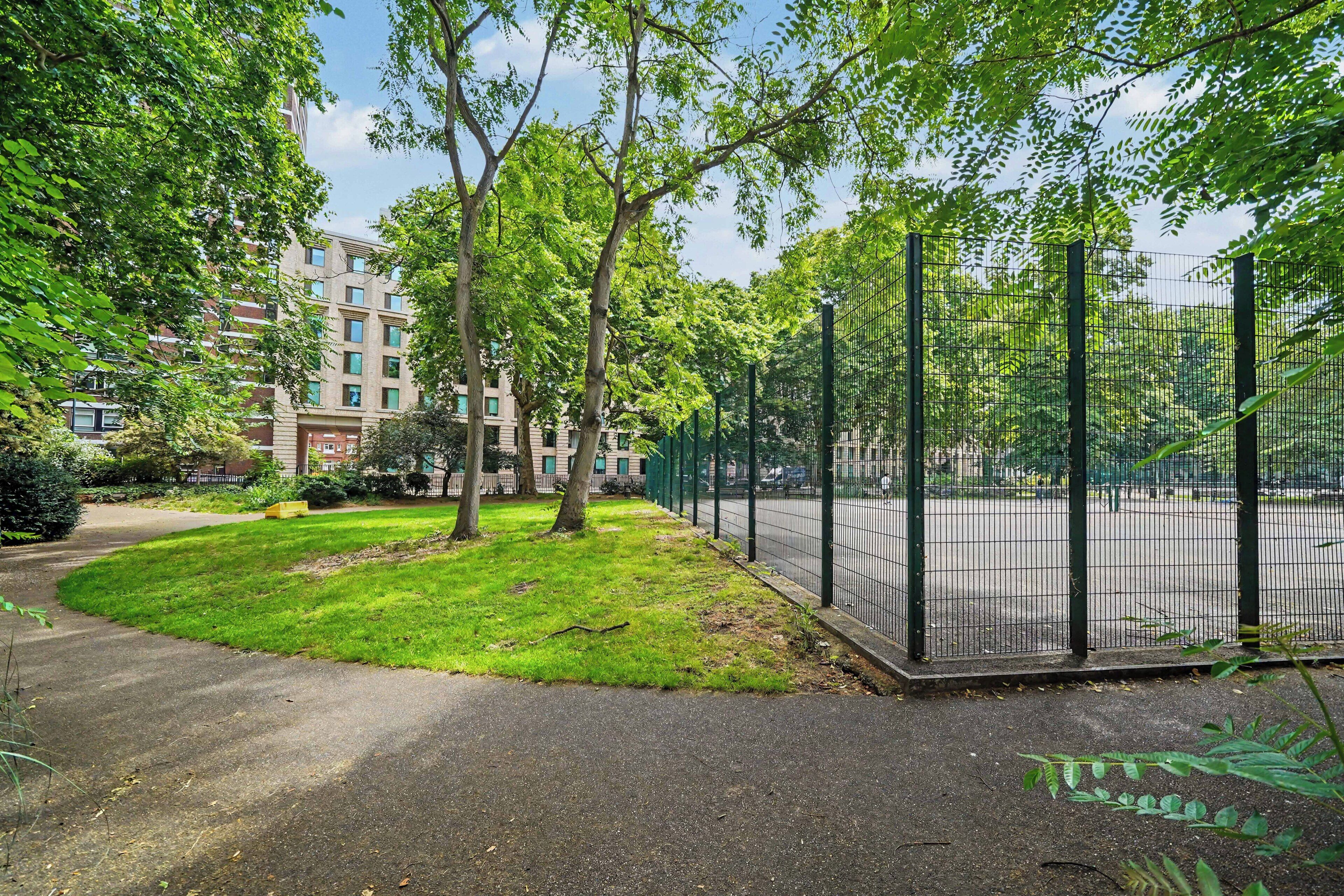A park with a fenced tennis court, trees, and a building in the background.