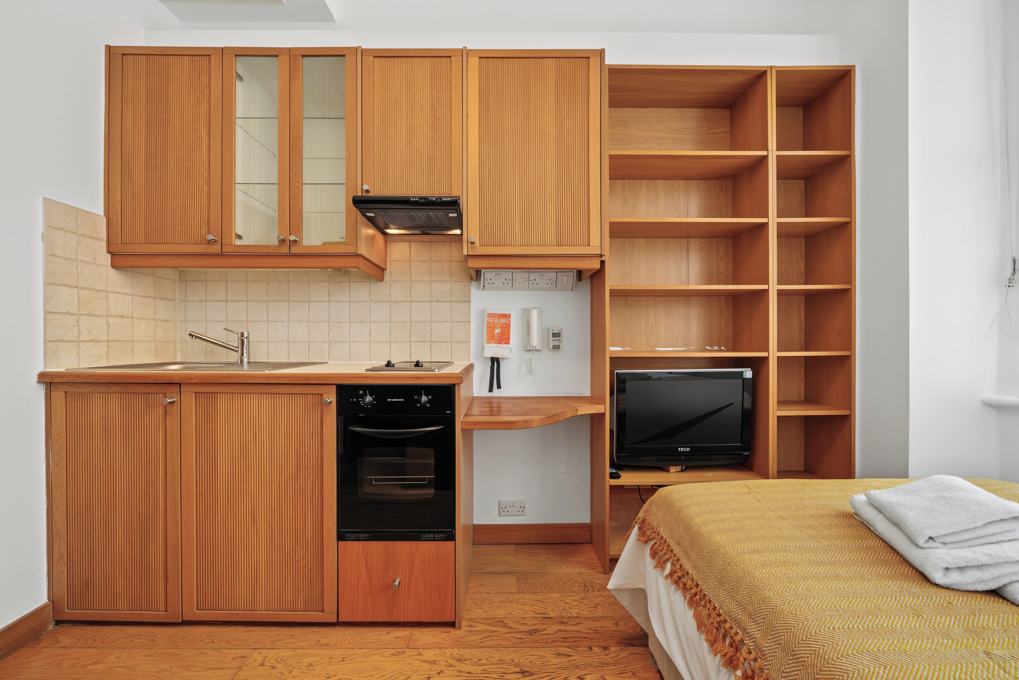 A compact kitchen with wooden cabinets, a built-in oven, a refrigerator, and a television on a wooden shelf.