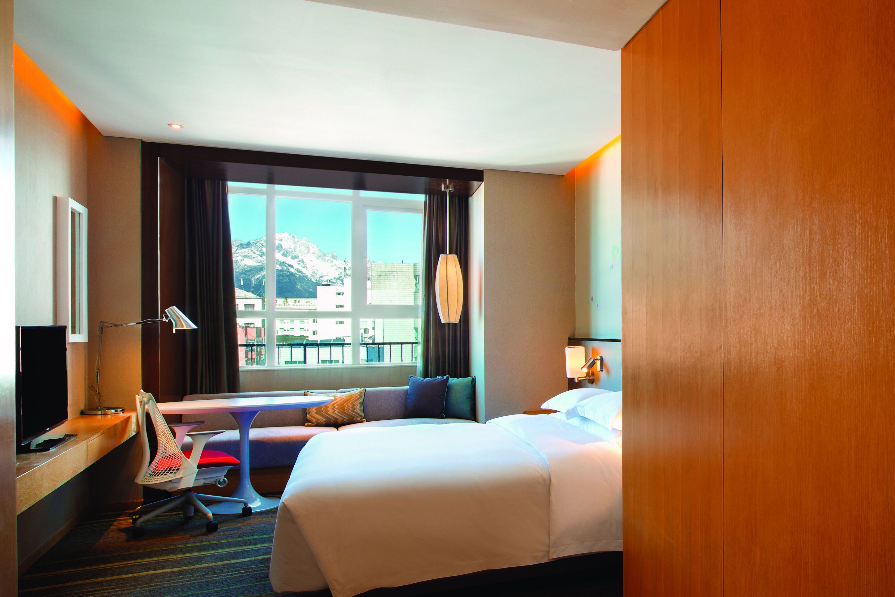 Une chambre d’hôtel équipée d’un lit, d’un bureau, d’une chaise, d’une télévision, d’une lampe et offrant une vue sur la montagne par la fenêtre.