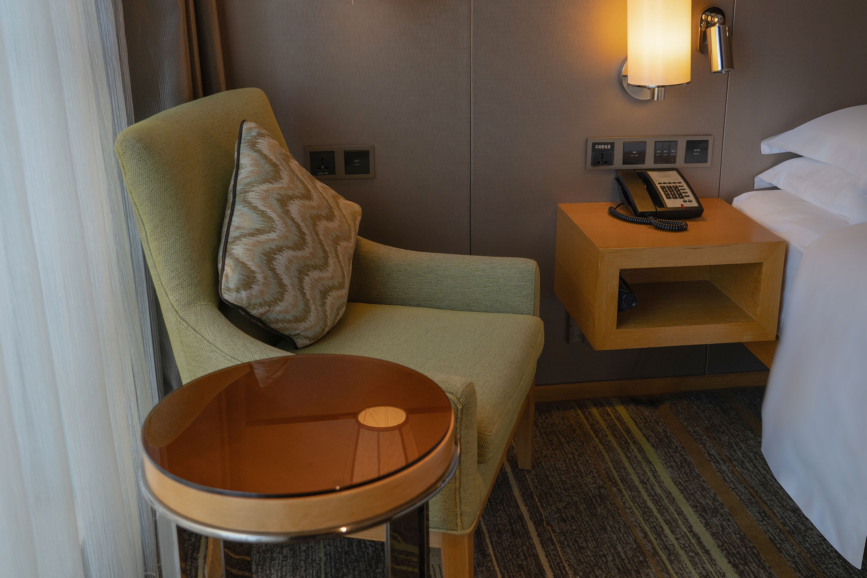 A hotel room with a green armchair, a small table, a bedside table with a lamp, a telephone, and a bed with white linens.
