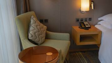 A hotel room with a green armchair, a small table, a bedside table with a lamp, a telephone, and a bed with white linens.