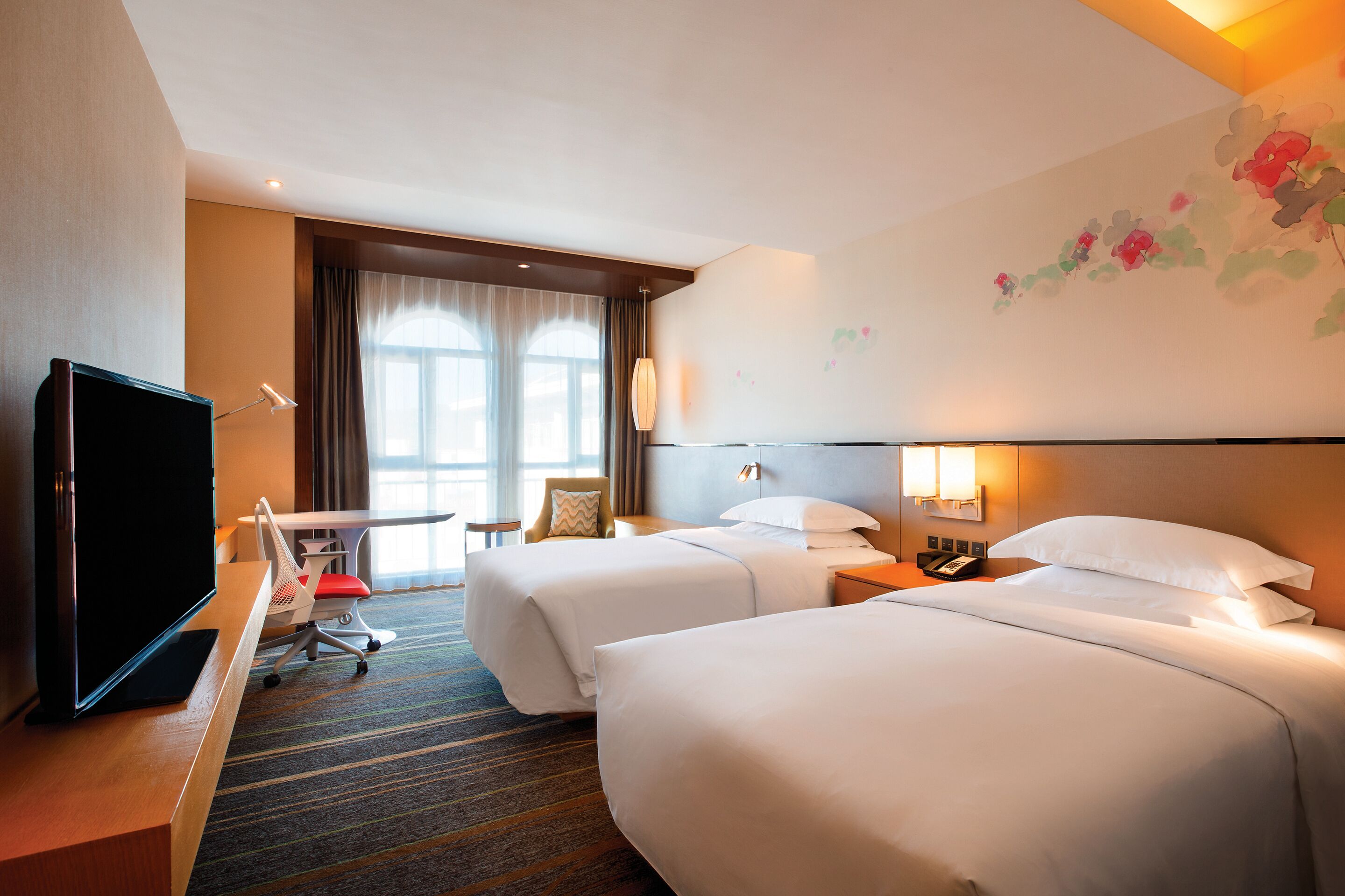 A hotel room with two beds, a desk with a TV, a chair, and a window with curtains.