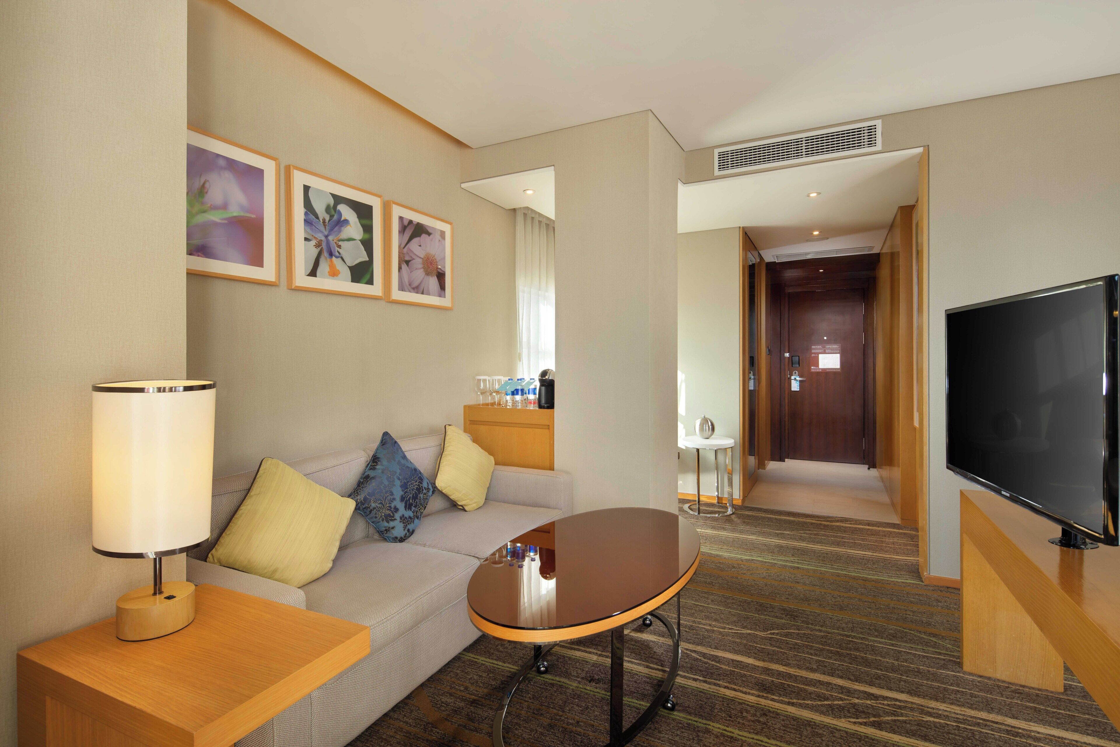 A hotel room with a sofa, a table, a lamp, a television, and framed pictures on the wall.