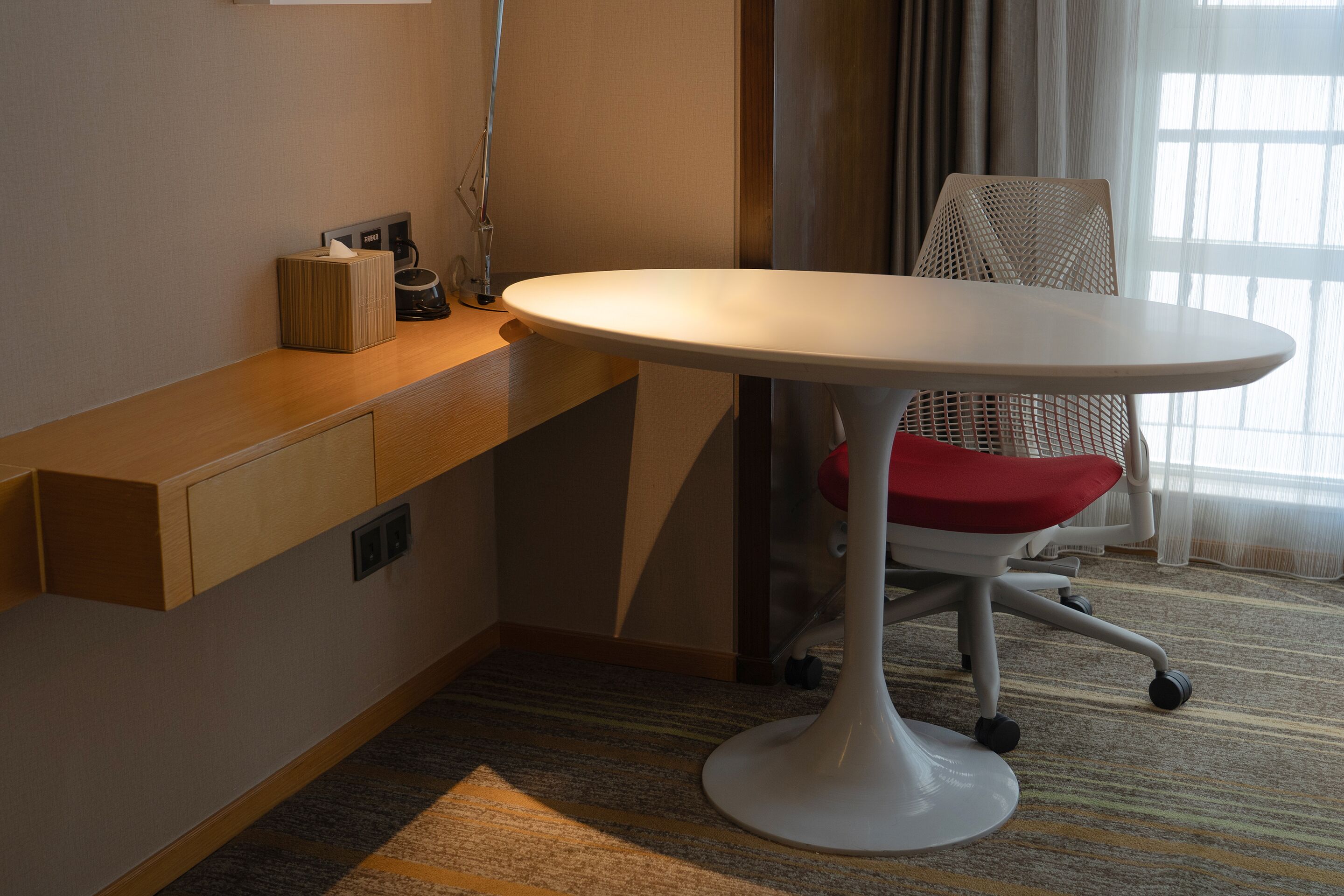 A hotel room with a white round table, a red cushioned chair, a wooden side table, and a patterned carpet.