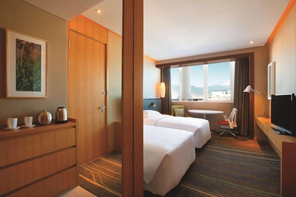 A hotel room with two beds, a desk with a TV, a small table, and a window with a city view.