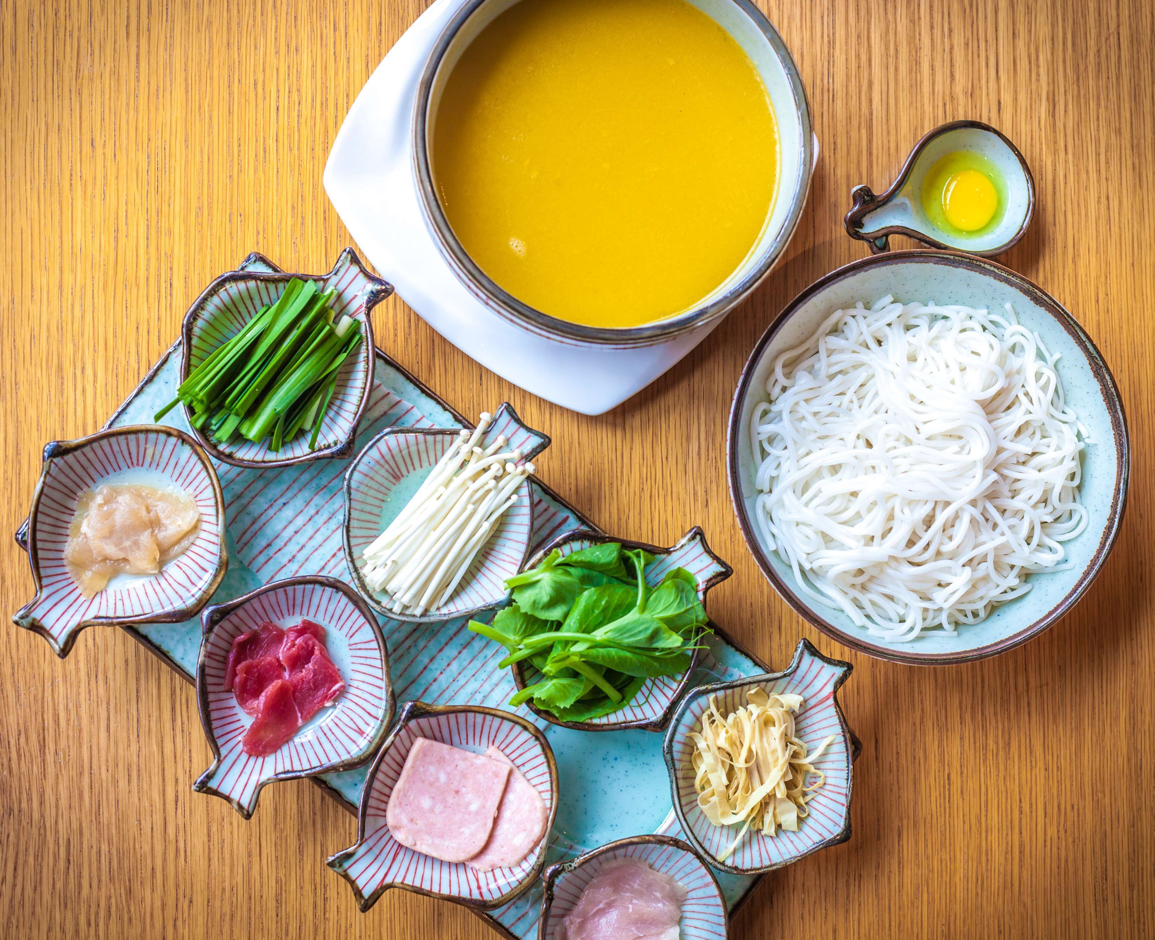 A table with various food items including a bowl of soup, a bowl of noodles, and several small dishes containing different ingredients.