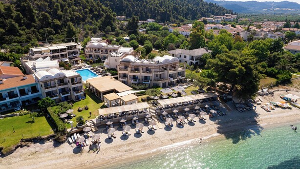 Vista aérea de un hotel costero con varias construcciones, piscina y zona de playa con sombrillas y tumbonas.