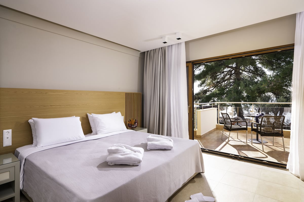 A hotel room with a large bed, a nightstand, a balcony with chairs, and a view of trees.