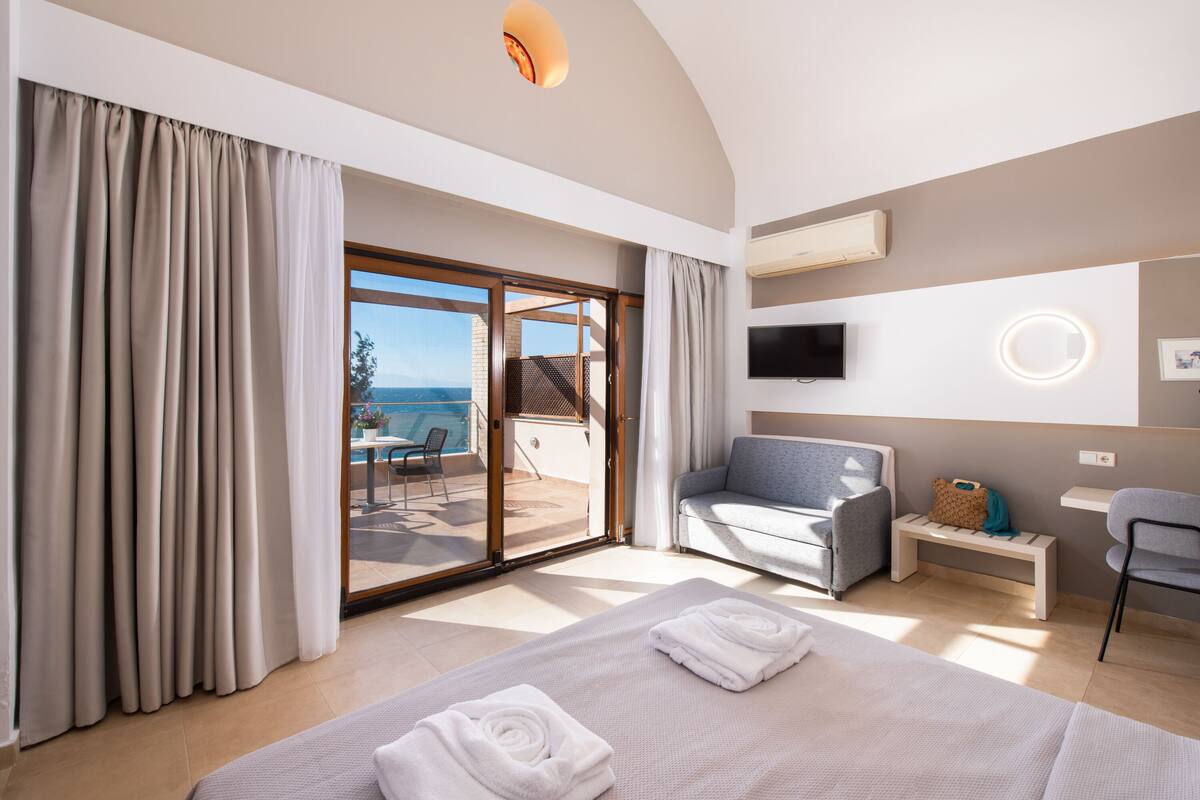 A modern hotel room with a bed, a sofa, a TV, and a view of the sea.