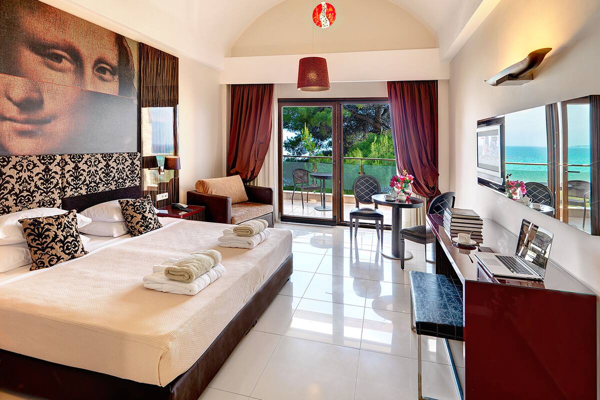 A spacious bedroom with a large bed, a desk with a laptop, a chair, and a balcony with a view of the sea.