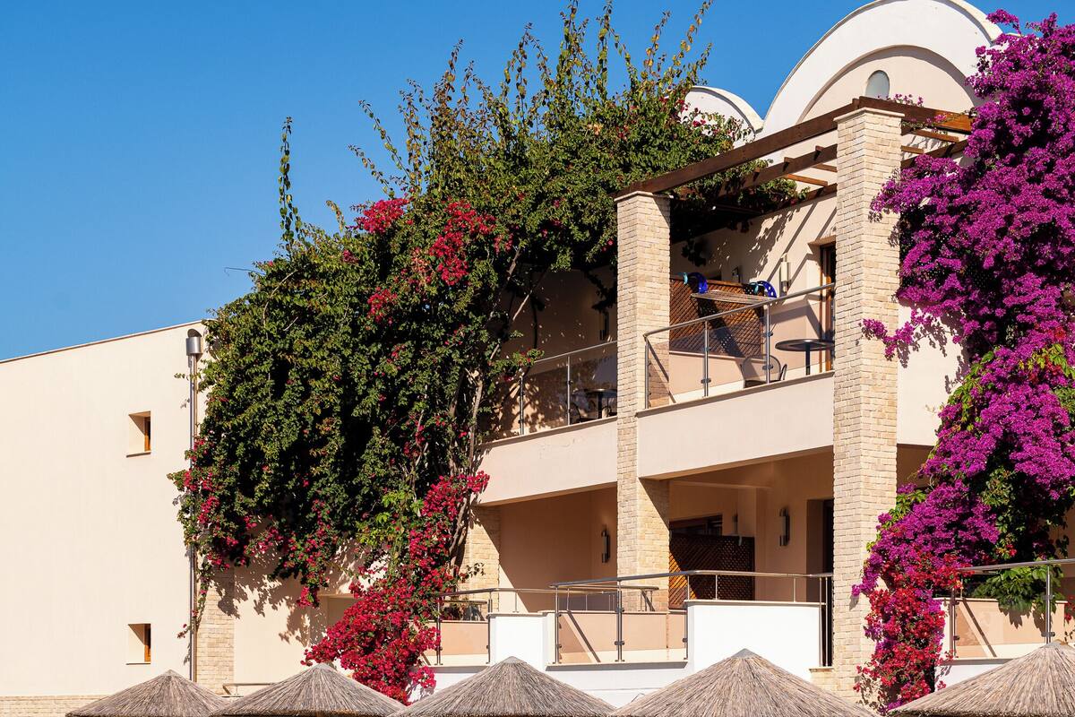 A swimming pool with lounge chairs and umbrellas, surrounded by a building with balconies and flowering plants.