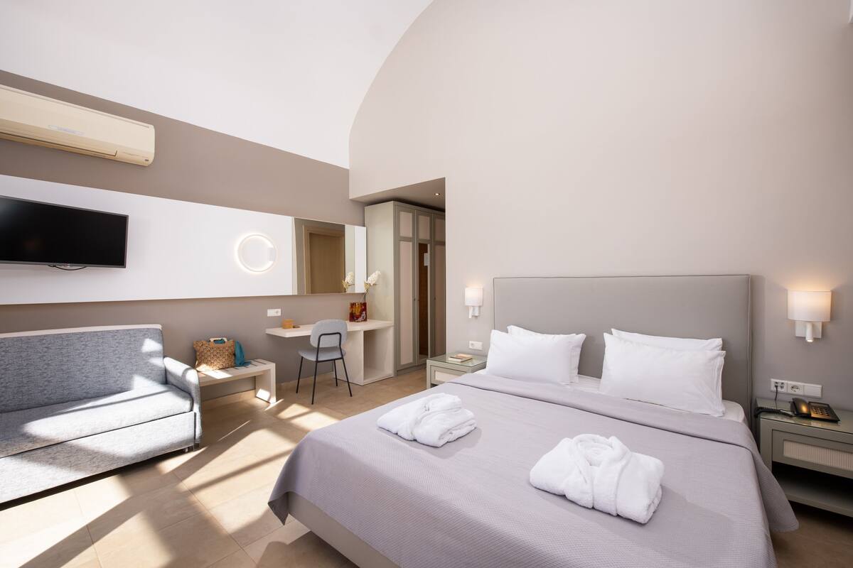 A modern hotel room with a large bed, a flat-screen TV, a desk with a chair, and a sofa.