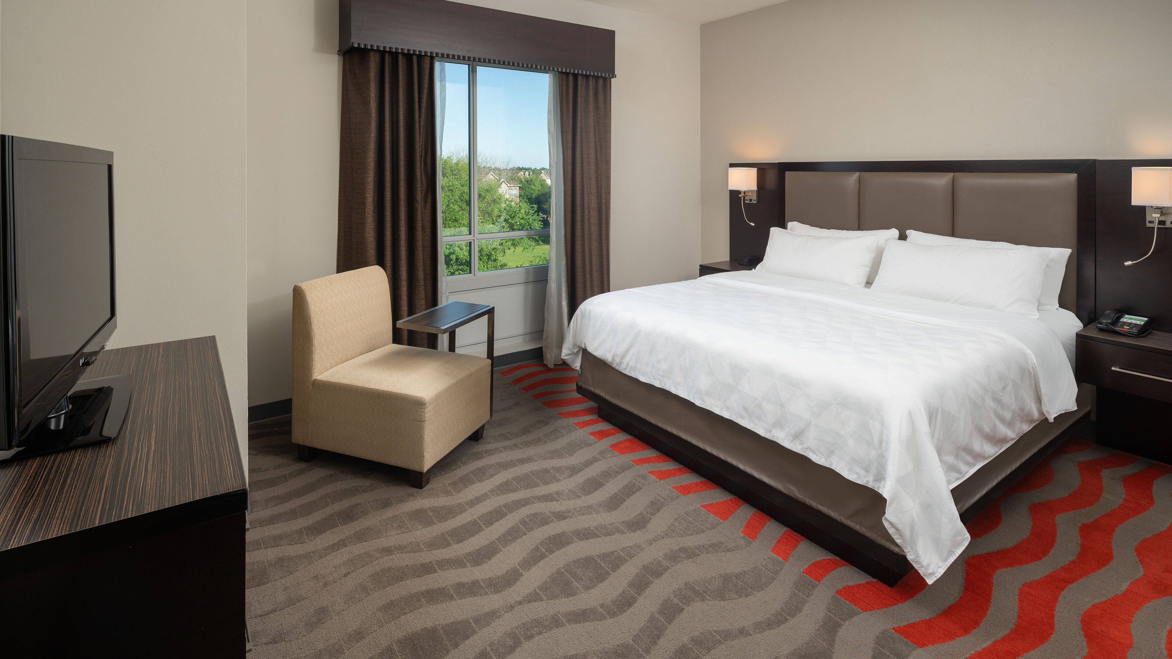 A hotel room with a bed, a chair, a TV, a small table, and a window with curtains.