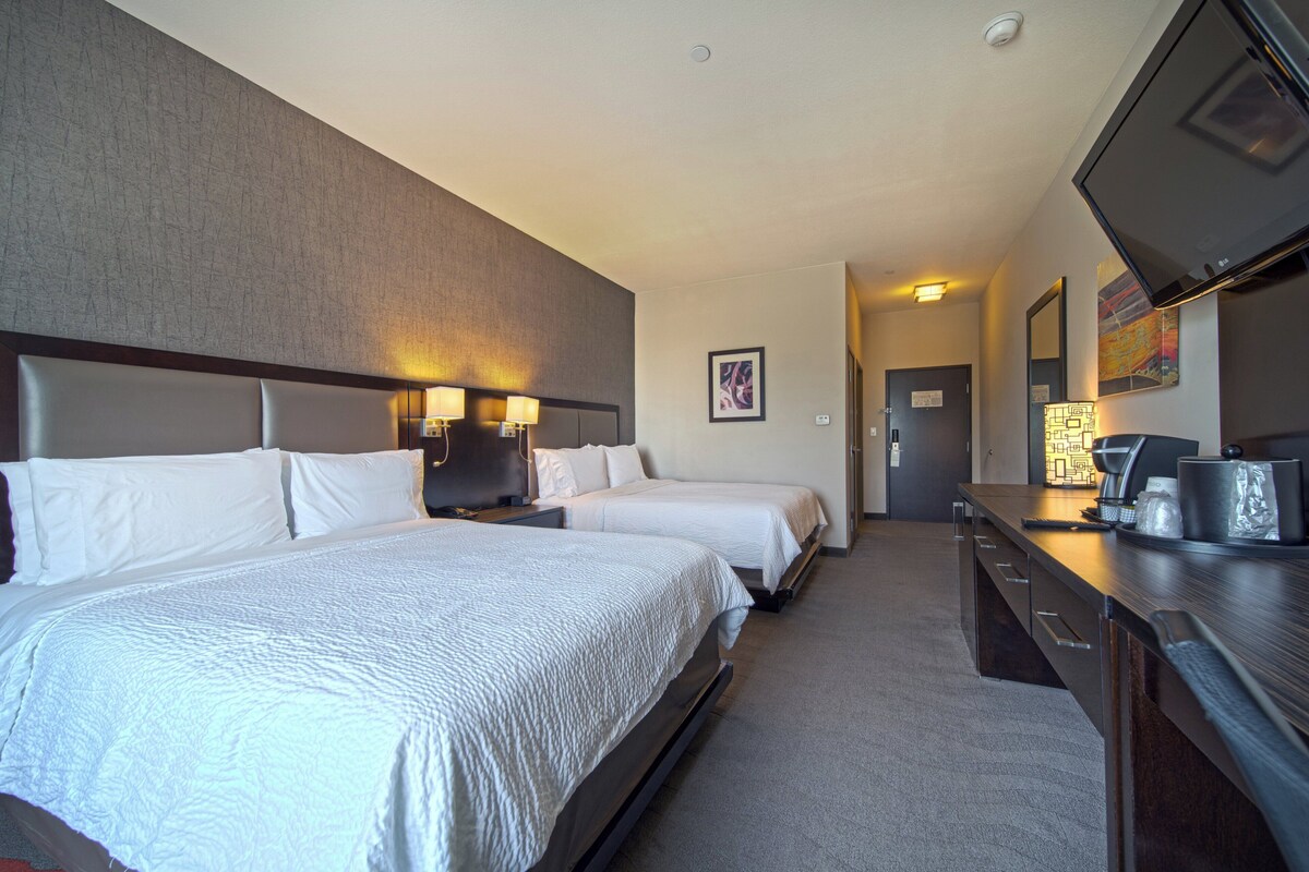 Ein Hotelzimmer mit zwei Betten, einem Schreibtisch, einem Flachbildfernseher und einer Badezimmertür im Hintergrund.