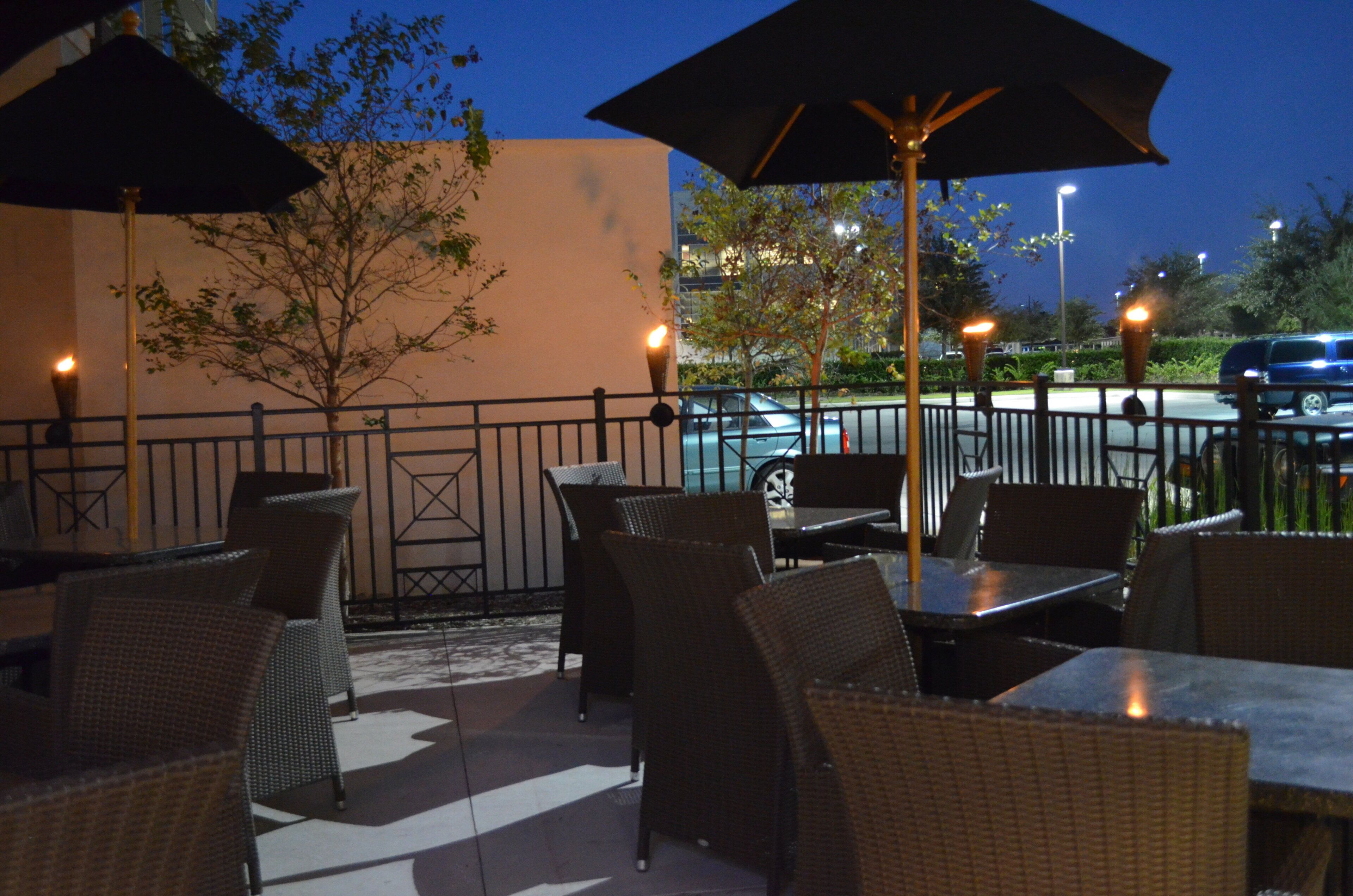 An outdoor patio with wicker furniture and umbrellas, a tree, and a street view in the background.