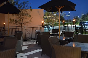An outdoor patio with wicker furniture and umbrellas, a tree, and a street view in the background. - Holiday Inn & Suites Houston West - Westway Park by IHG (Houston)
