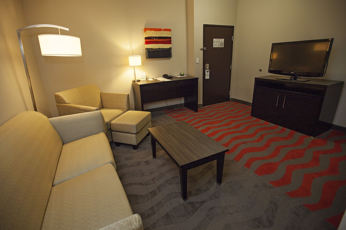 A hotel room with a sofa, armchair, desk, lamp, television, and a patterned carpet.