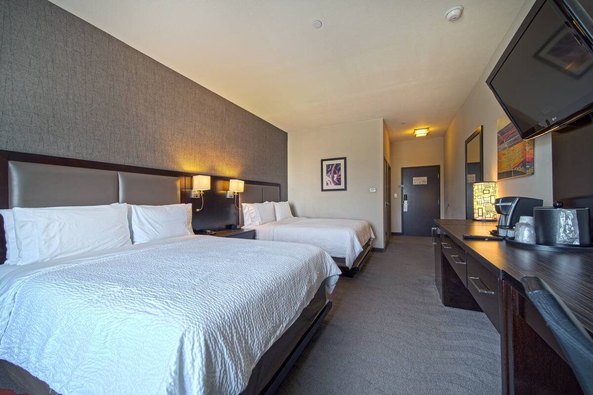 A hotel room with two beds, a desk, a flat-screen TV, and a bathroom door in the background.