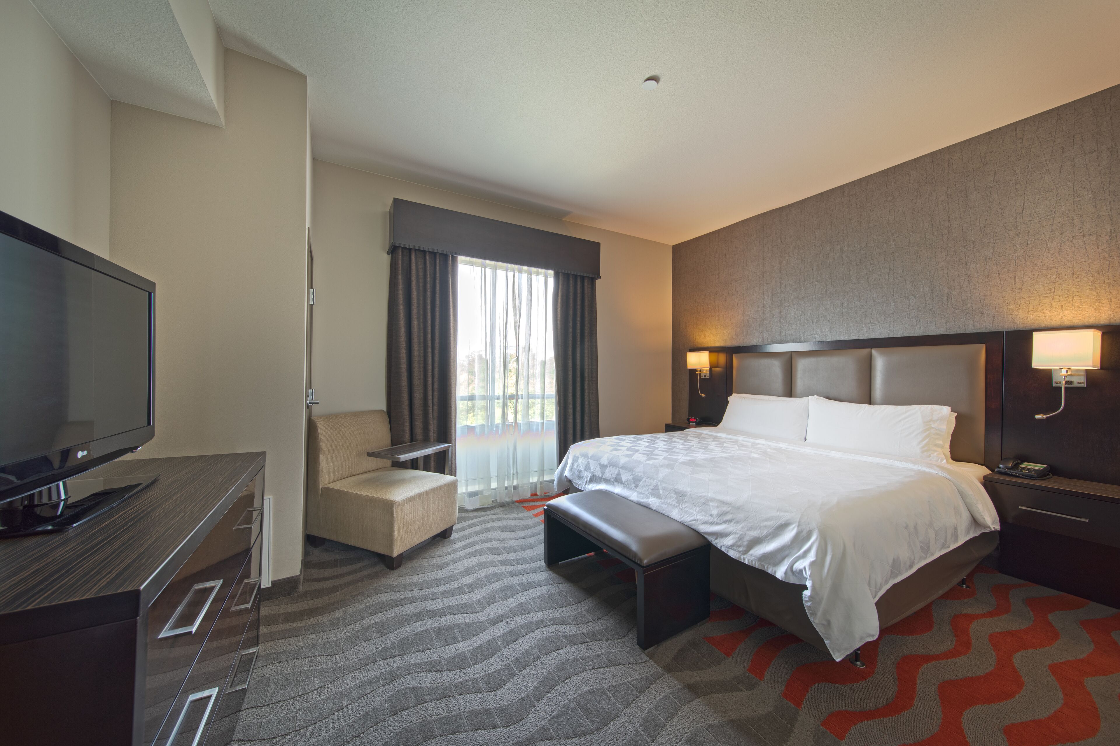 A hotel room with a bed, a desk with a TV, a chair, a bench, and a window with curtains.