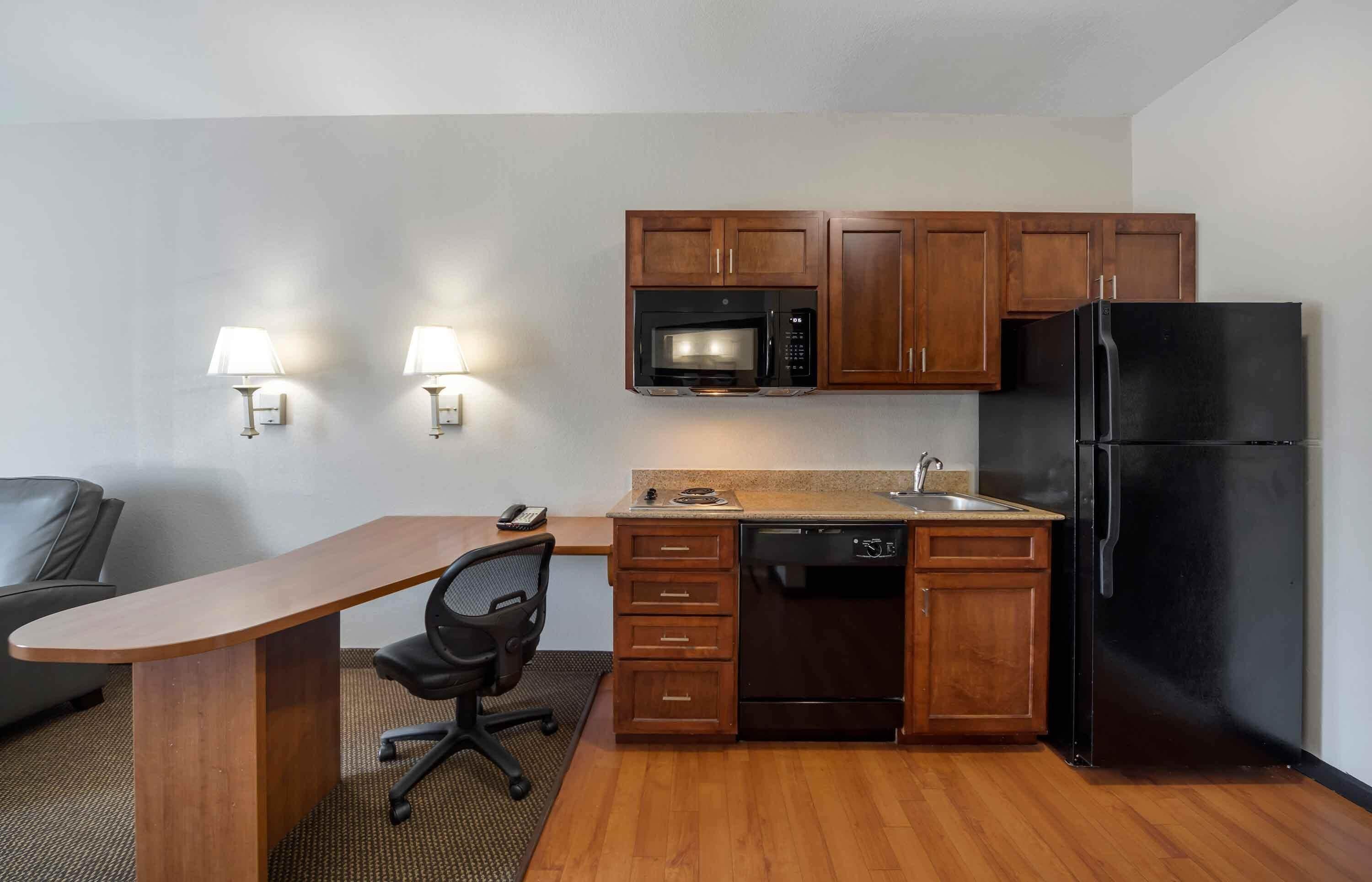A compact kitchen with wooden cabinets, a microwave, a refrigerator, and a small dining area with a chair and a desk.