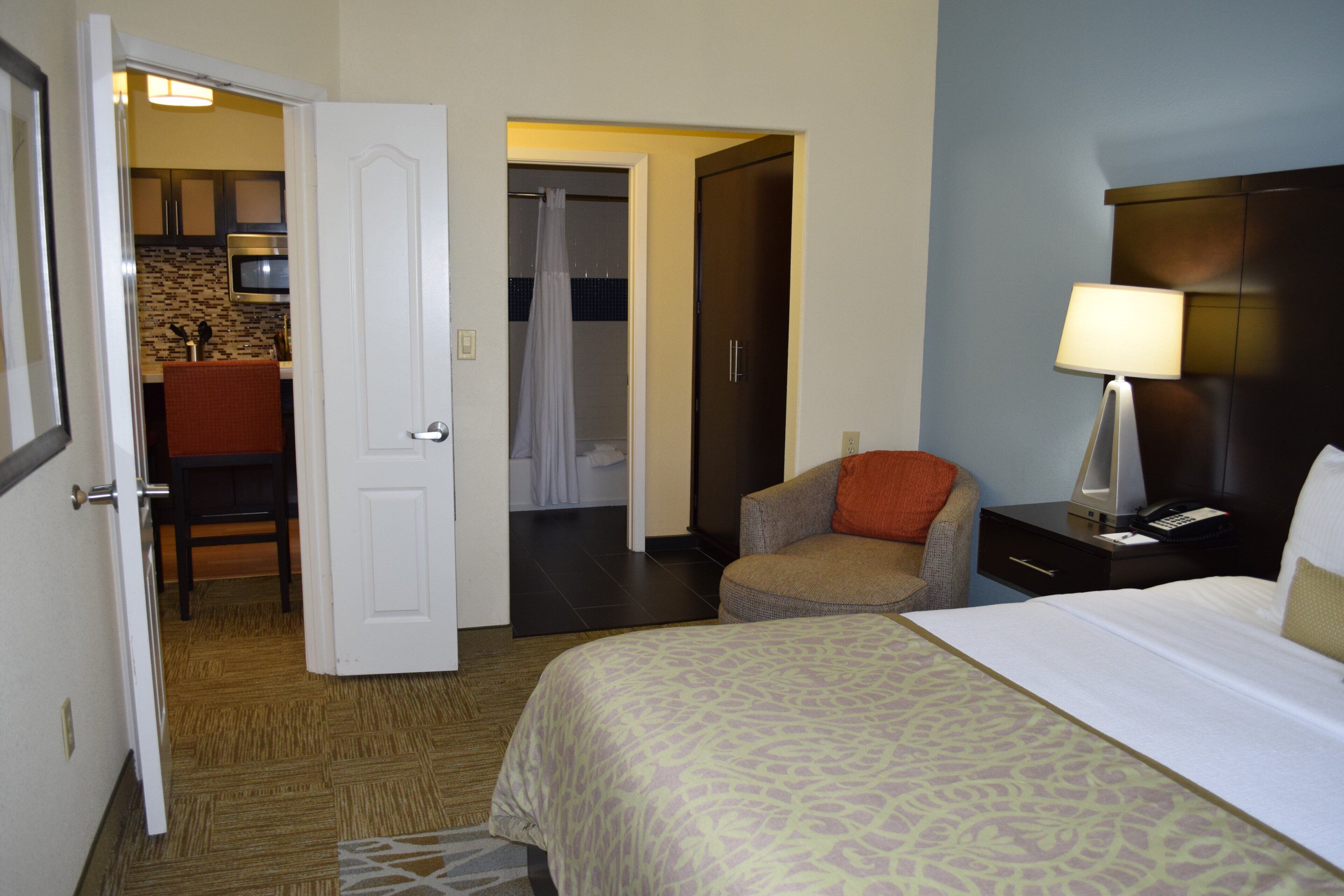 Une chambre d’hôtel avec un lit, une chaise, un bureau avec une lampe et une salle de bain visible à travers une porte ouverte.