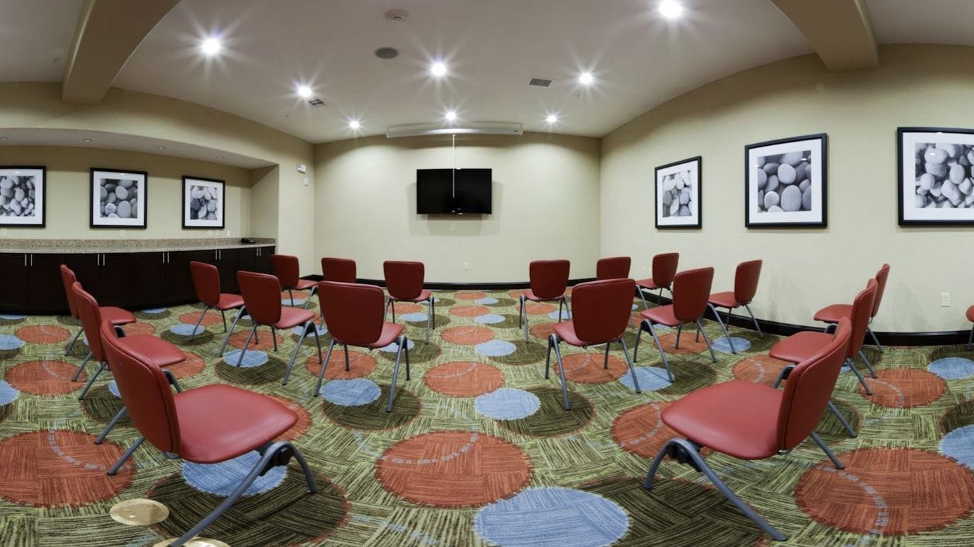 Un salón de conferencias con sillas rojas dispuestas en filas, un televisor de pantalla plana montado en la pared y alfombra estampada.