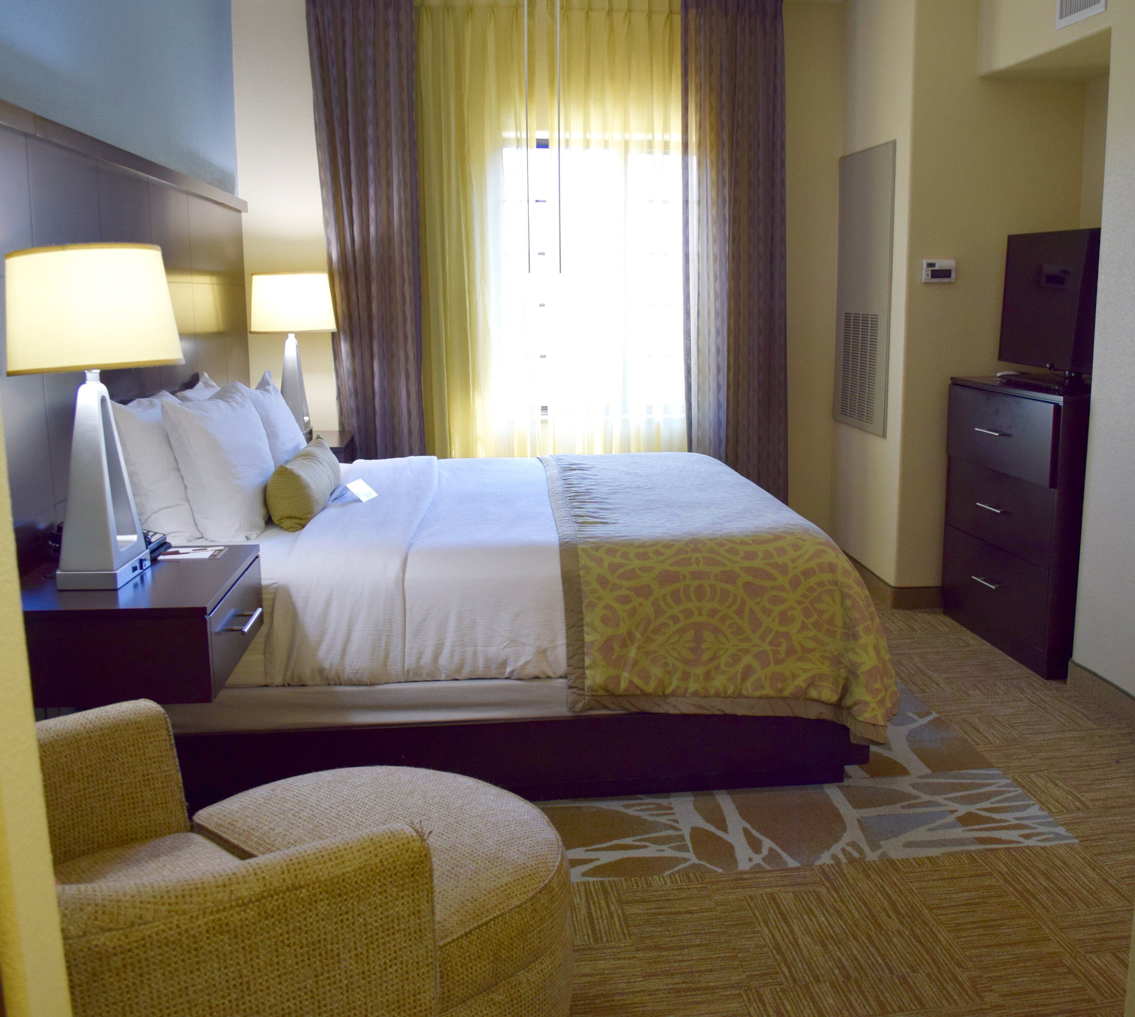 Una habitación de hotel con cama, escritorio con lámpara, una silla, cómoda y ventana con cortinas.