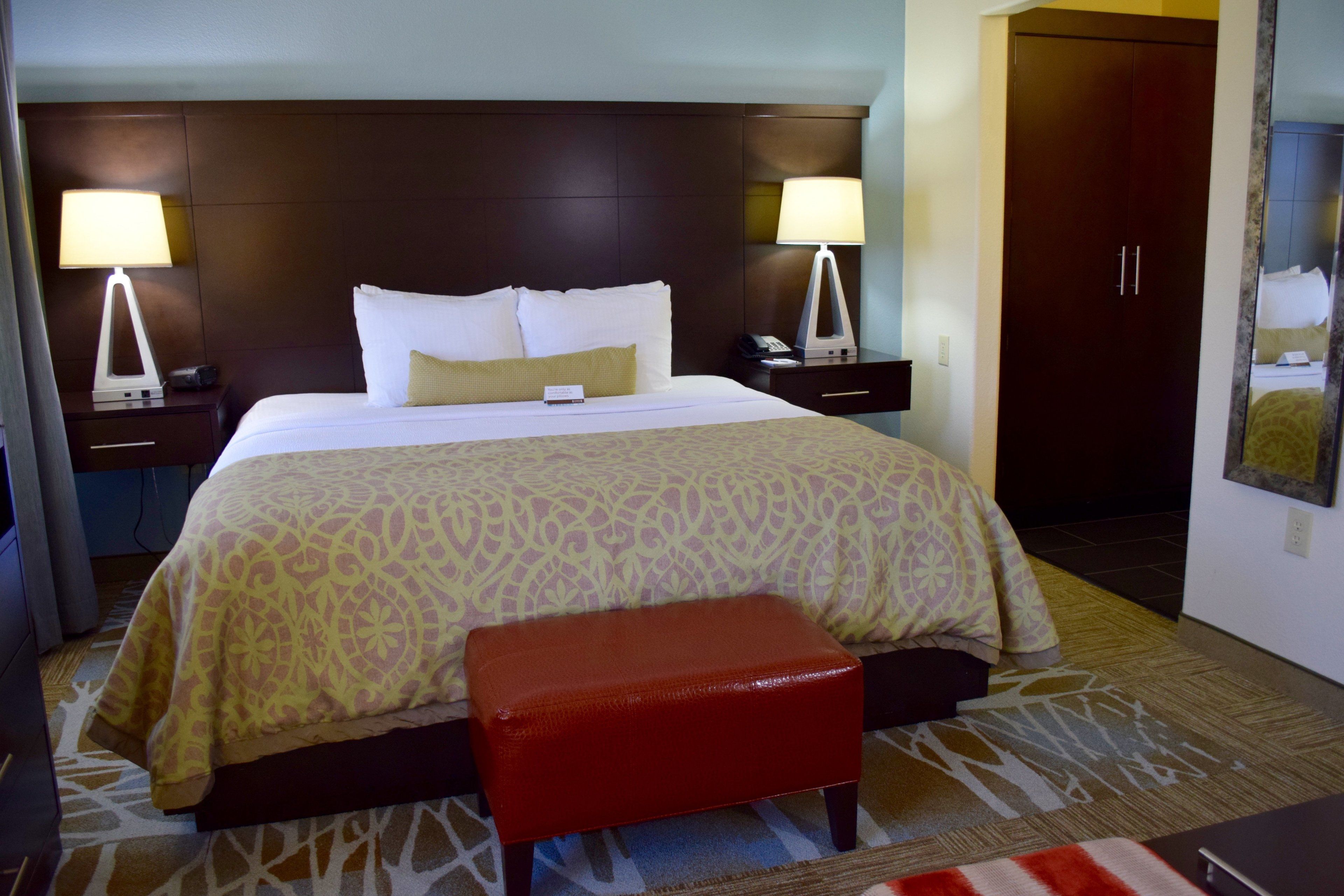 A hotel room with a large bed, two bedside lamps, a nightstand, a red bench, and a wardrobe.