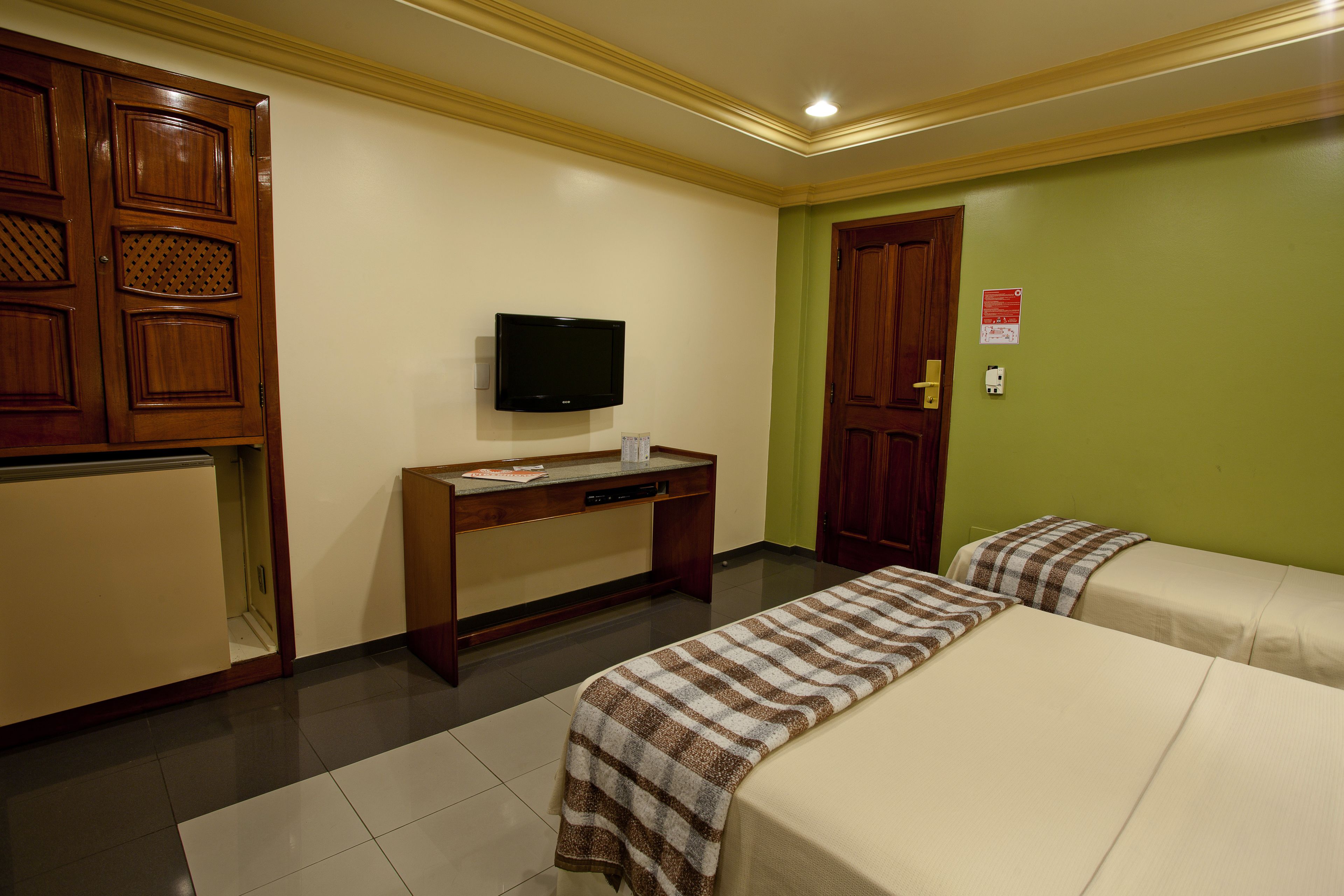 Quarto de hotel com duas camas, uma escrivaninha e uma televisão, e um balcão de recepção.