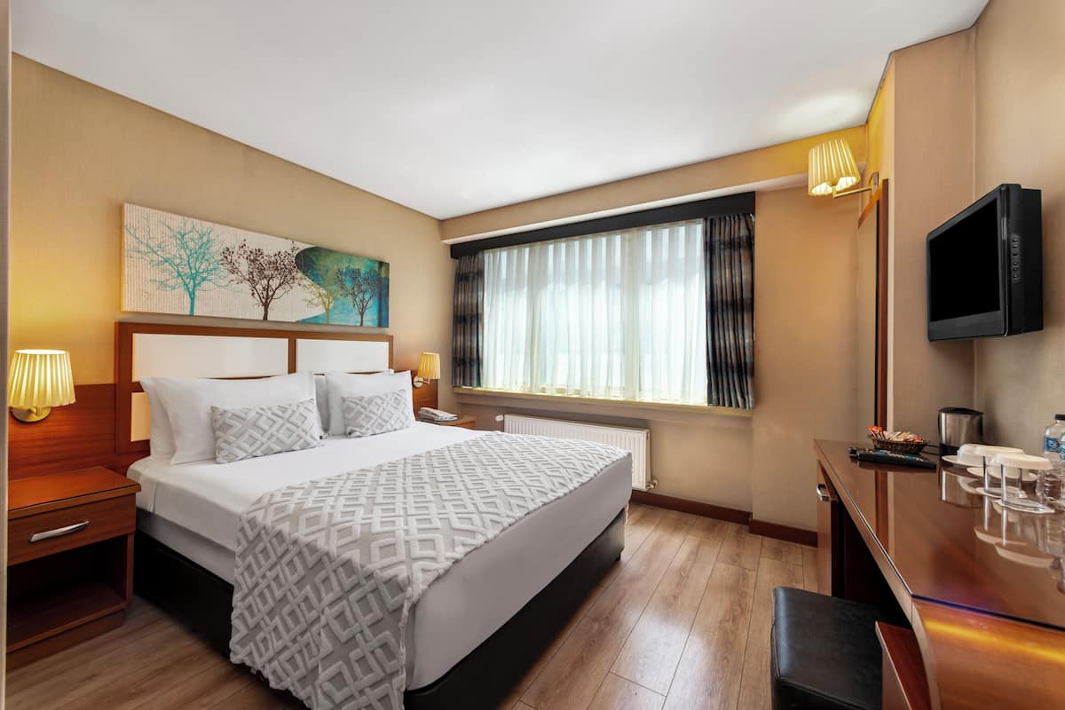Ein Hotelzimmer mit einem großen Bett, einem Schreibtisch mit Flachbildfernseher, einem Fenster mit Vorhängen und einem Bild an der Wand.