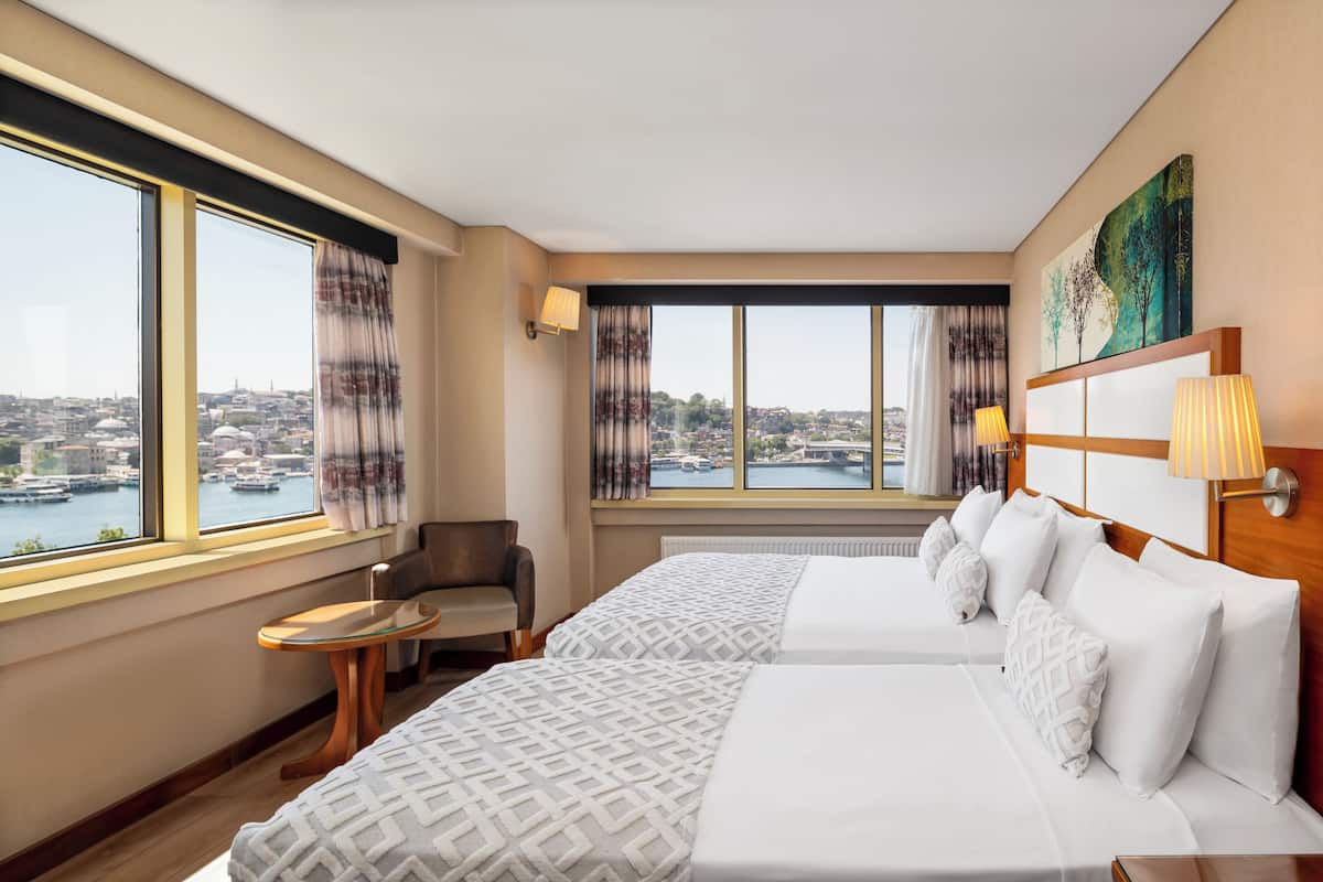 Ein Hotelzimmer mit zwei Betten, Blick auf eine Stadtkulisse durch große Fenster, einem kleinen Tisch und einem Stuhl.