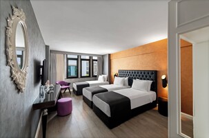 Habitación de hotel con tres camas, un espejo grande, un escritorio con silla y vistas a la ciudad a través de amplias ventanas.