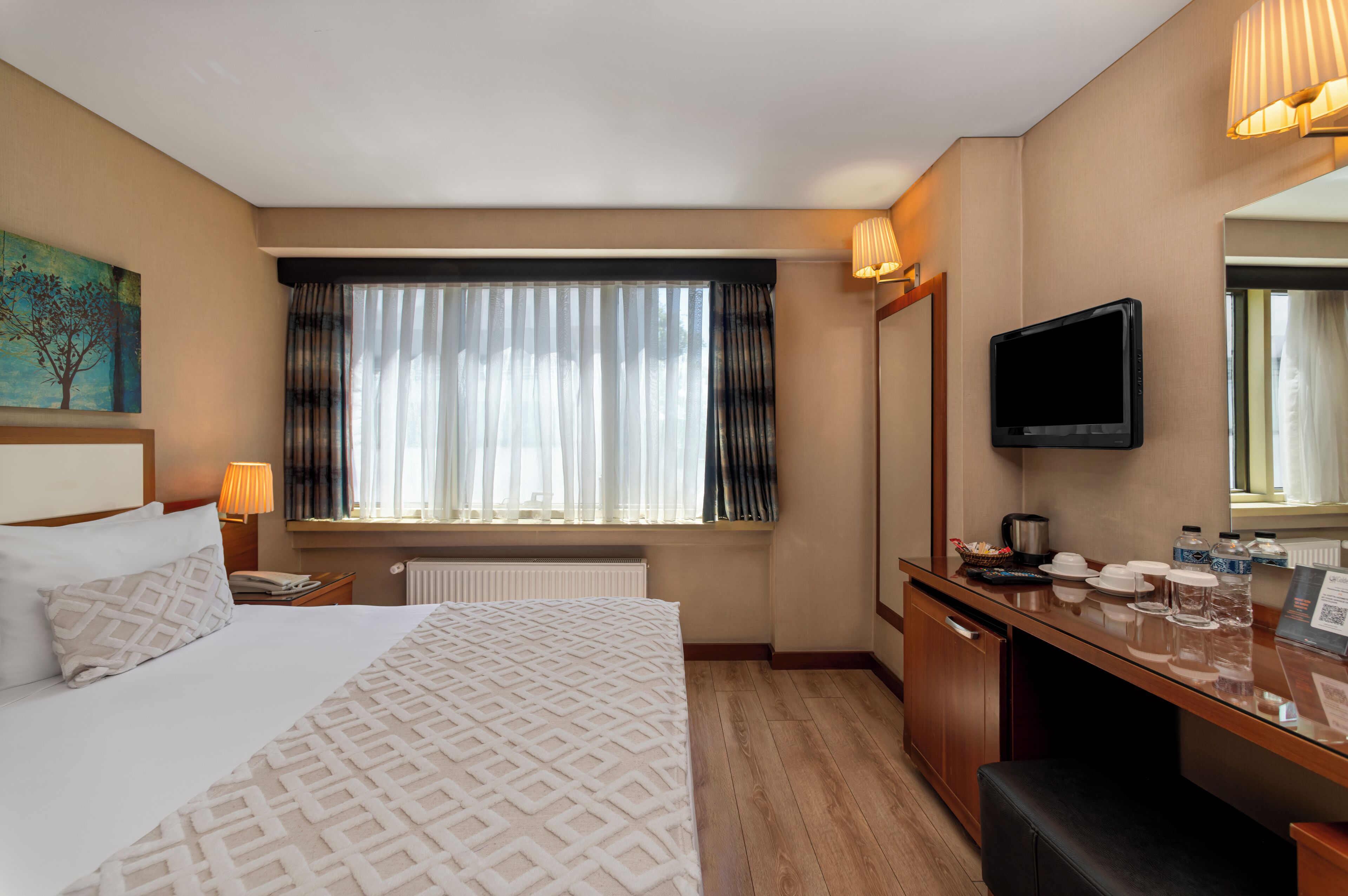 A hotel room with a large bed, a desk with a flat-screen TV, a window with curtains, and a painting on the wall.