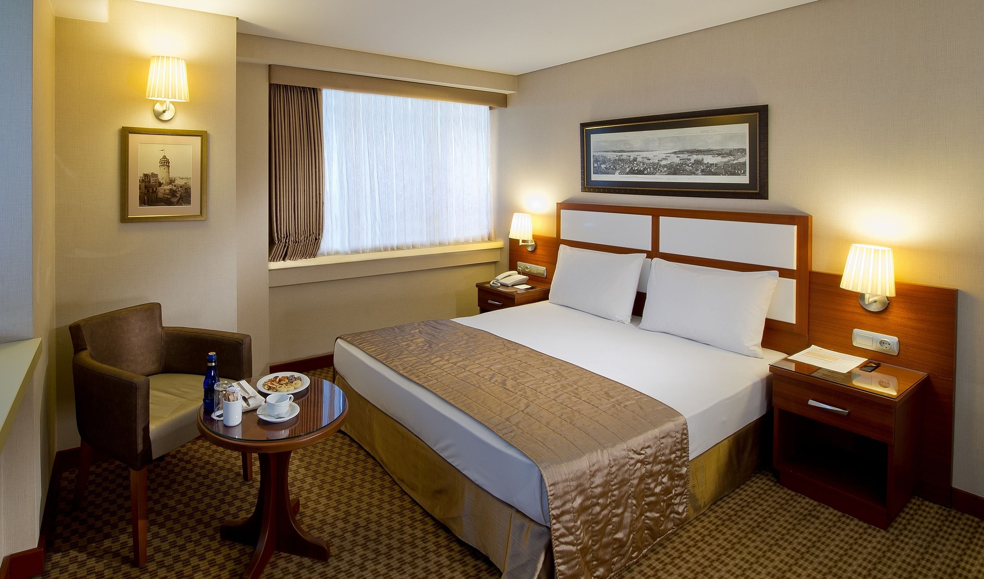 Ein Hotelzimmer mit Bett, Nachttischen, einem Sessel, einem kleinen Tisch mit Getränken und Snacks, einem Fenster mit Vorhängen und Wandleuchten.