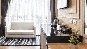 Een moderne hotelkamer met een bank, een glazen salontafel, een flatscreen televisie aan de muur en uitzicht op de stad door grote ramen.