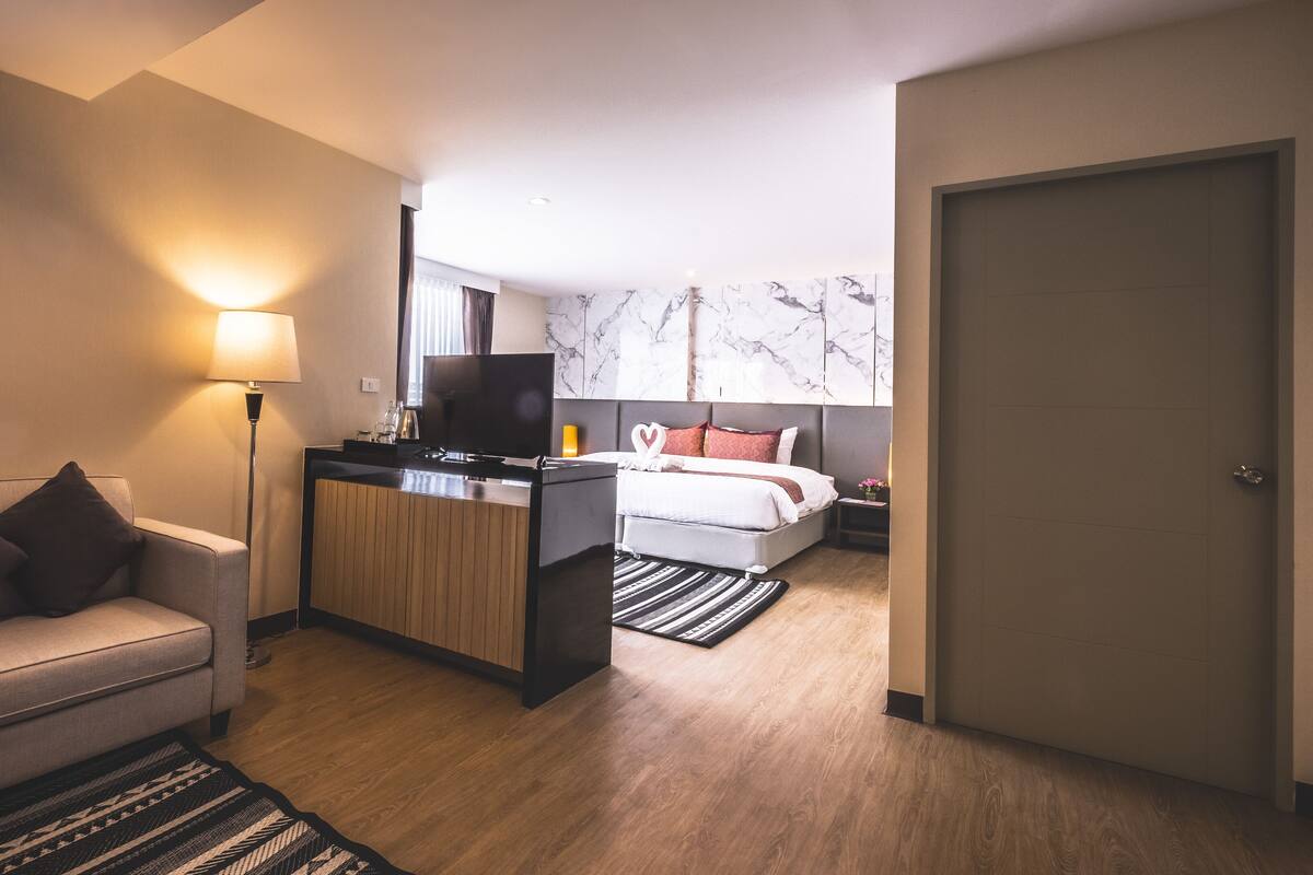 A modern hotel room with a sofa, a desk with a flat-screen TV, a bed with a headboard, and a marble wall.