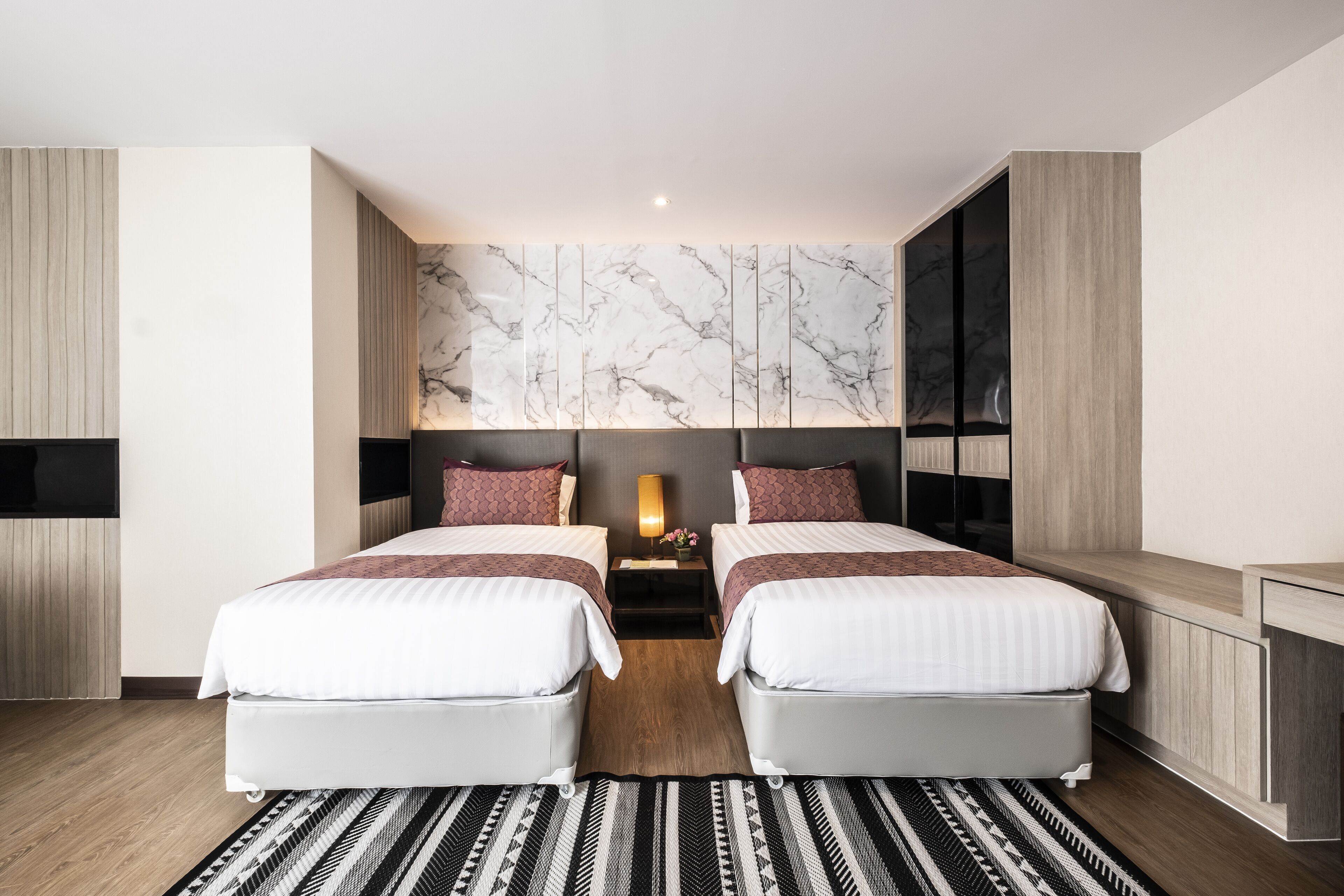A modern hotel room with two beds, a wooden floor, a marble-patterned wall, and a small bedside table with a lamp.