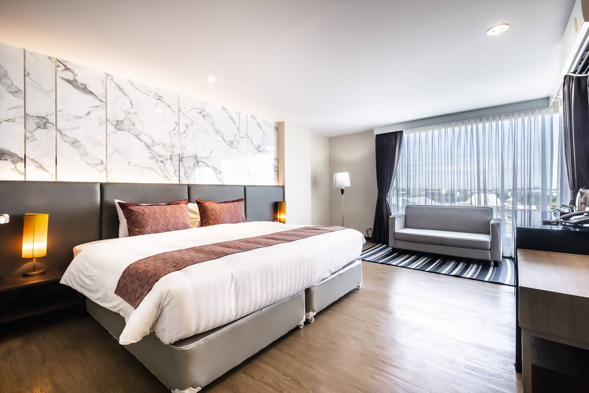 A modern hotel room with a large bed, a sofa, a desk, and a view of the city.