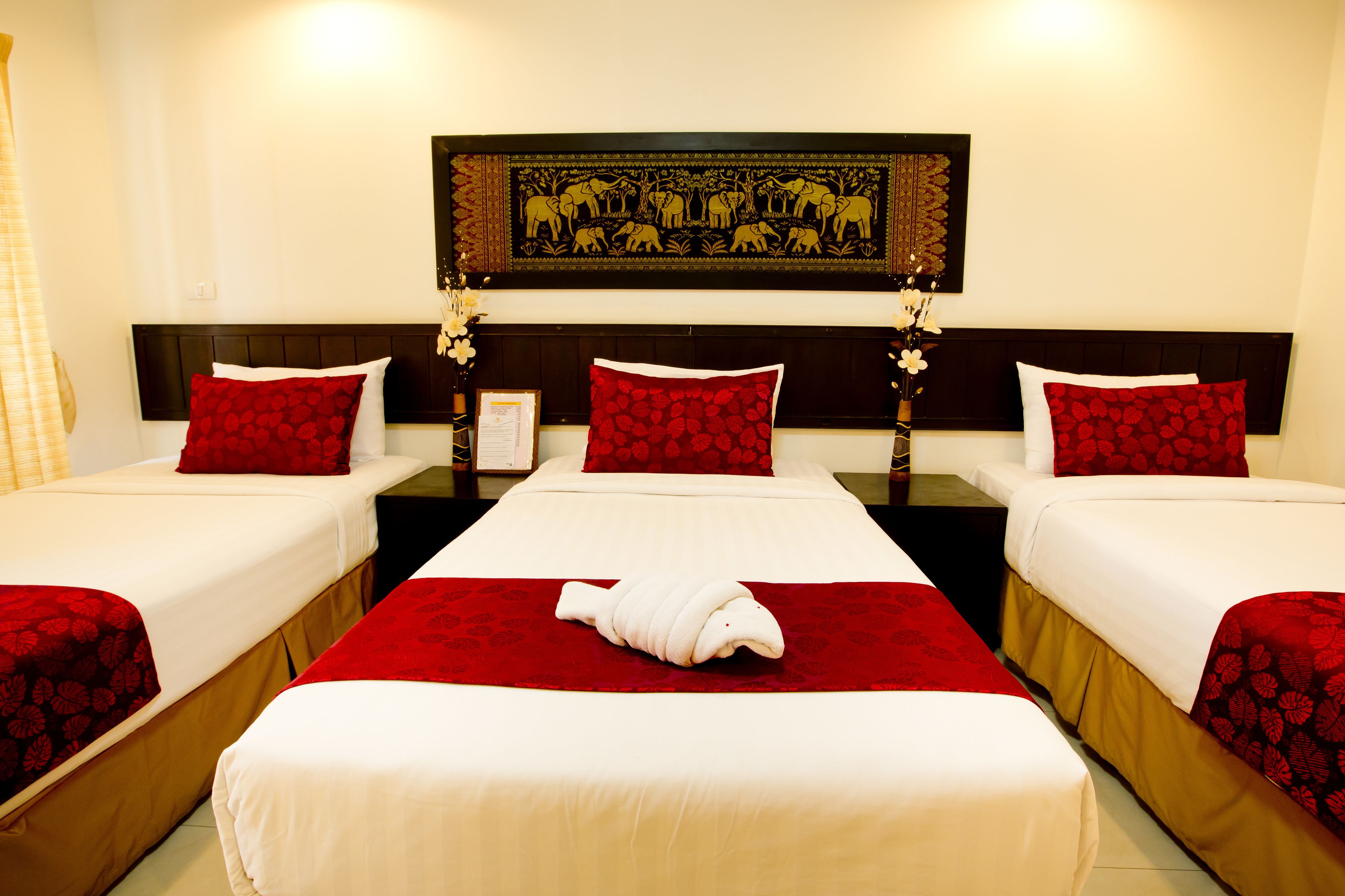 Quarto de hotel com três camas, cada uma com roupa de cama estampada em flores vermelhas, uma cama com toalha e um adorno decorativo na parede.